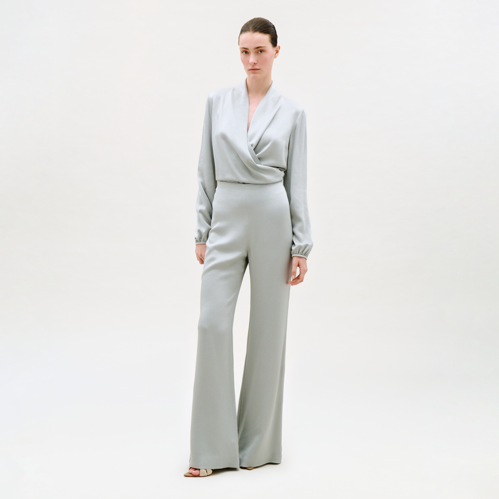 A woman with her hair pulled back and a neutral expression stands against a plain light background, wearing the Palermo Pant—light gray, wide-leg style—paired with a matching long-sleeve wrap-front top and open-toe heels.