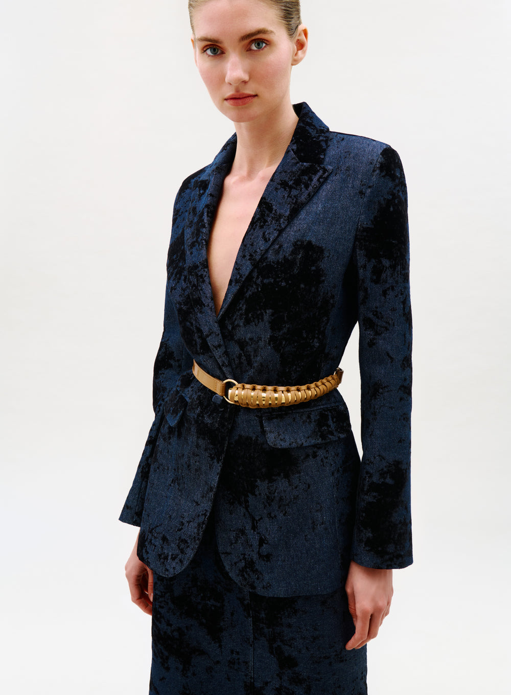 A woman with light skin and blue eyes stands against a plain background wearing the Rebeca Jacket, a dark blue textured velvet blazer paired with a gold woven waist belt and matching skirt; her hair is pulled back.