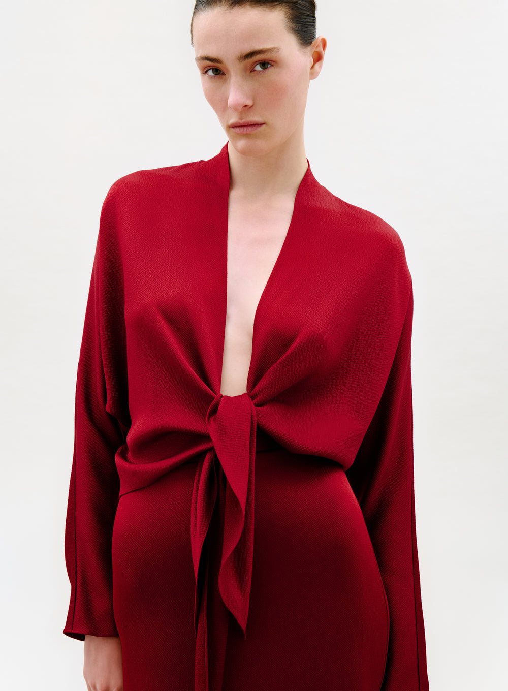 A woman with dark hair pulled back wears the deep red Reiko Blouse featuring long sleeves, an open front, and a large knotted waist tie, standing against a plain white background with a neutral expression.