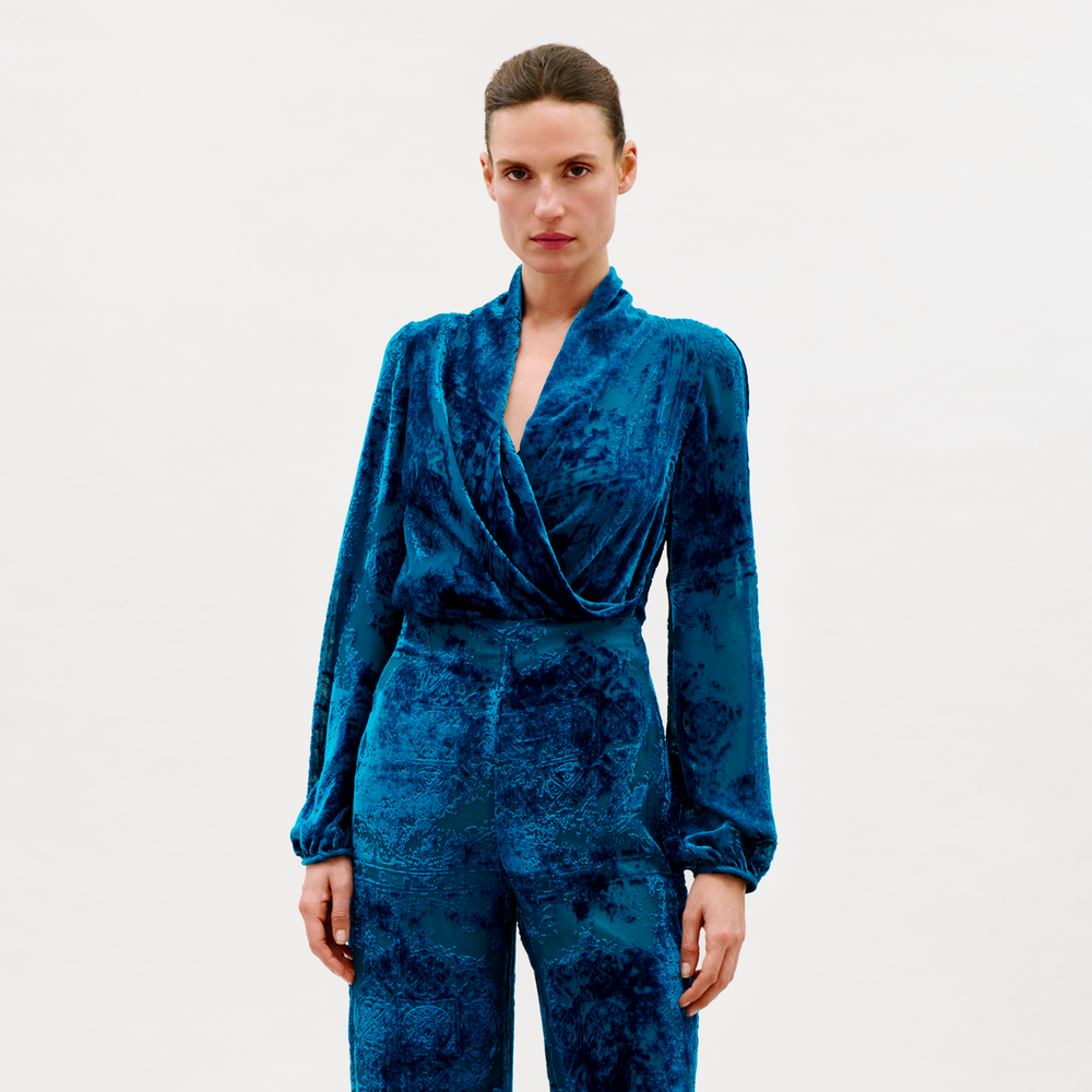 A woman stands against a plain light background, wearing the Tea Blouse in blue velvet with matching high-waisted pants. She looks directly at the camera with a neutral expression.