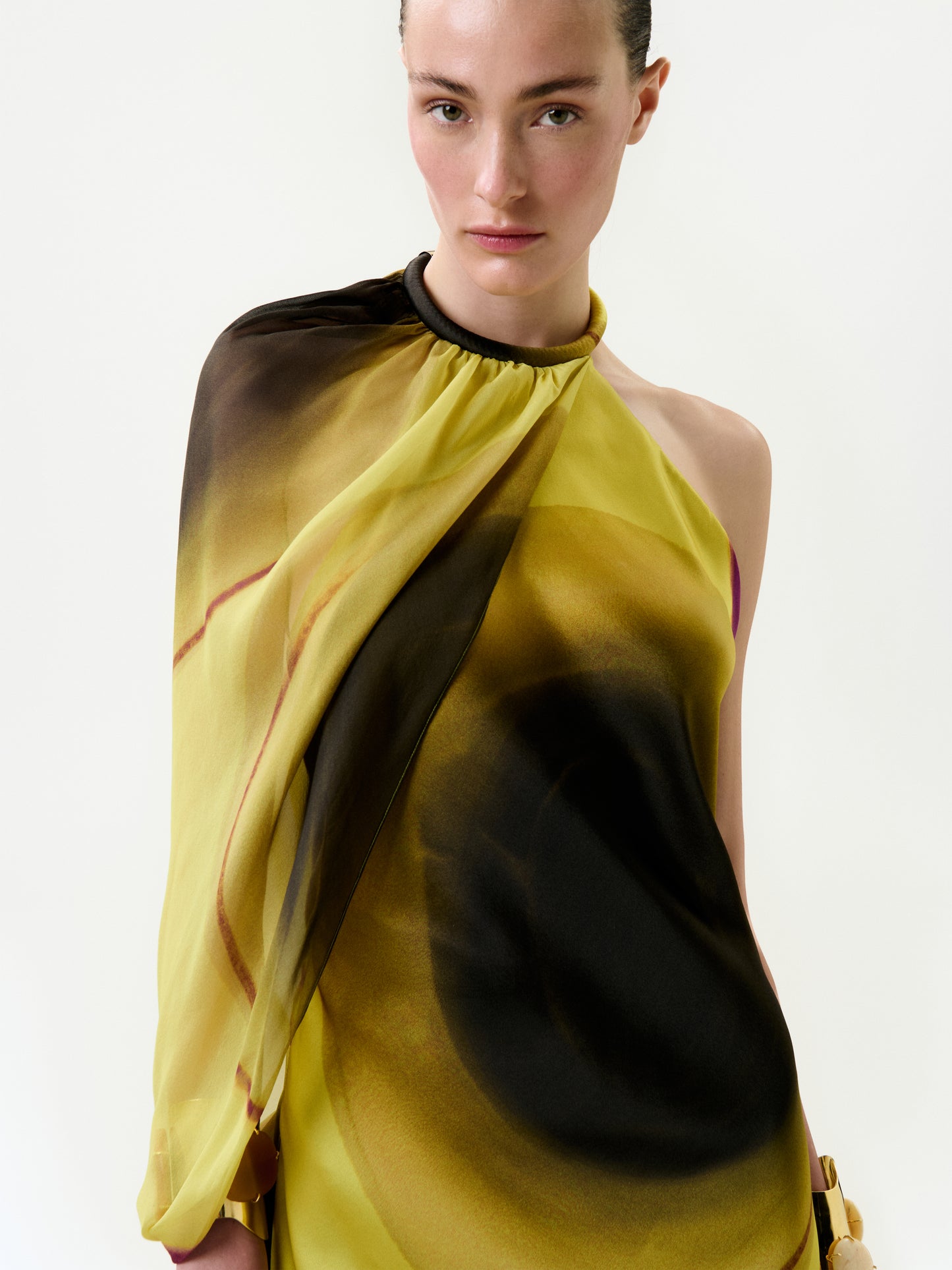 A woman with light skin and tied-back hair wears the Mari Luz Dress, a yellow and brown one-shoulder piece made from luxurious Italian fabric with a circular pattern, standing against a plain white background.