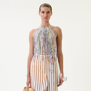 A woman wears the Lianne Top, a halter style with colorful fringe, paired with a beige-and-white vertical striped skirt. She holds a small handbag and a clear bracelet against a plain white background.