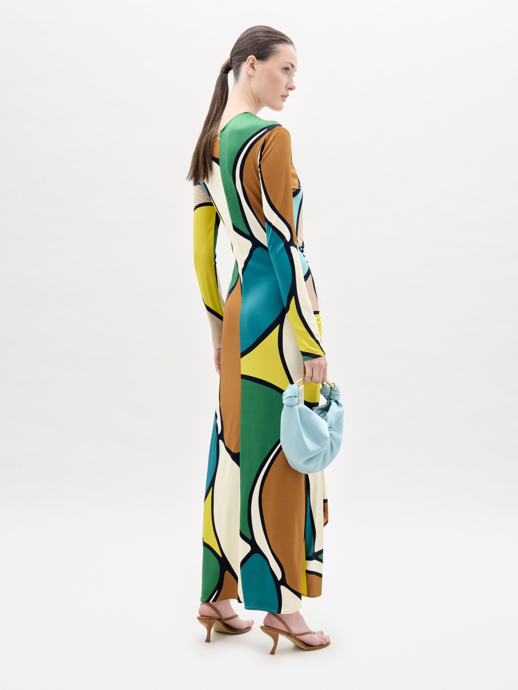 A woman stands against a white background wearing the Ilania Dress, a long midi with an abstract pattern in yellow, green, blue, brown, and black Italian fabric. She holds a small blue bag and wears brown sandals.