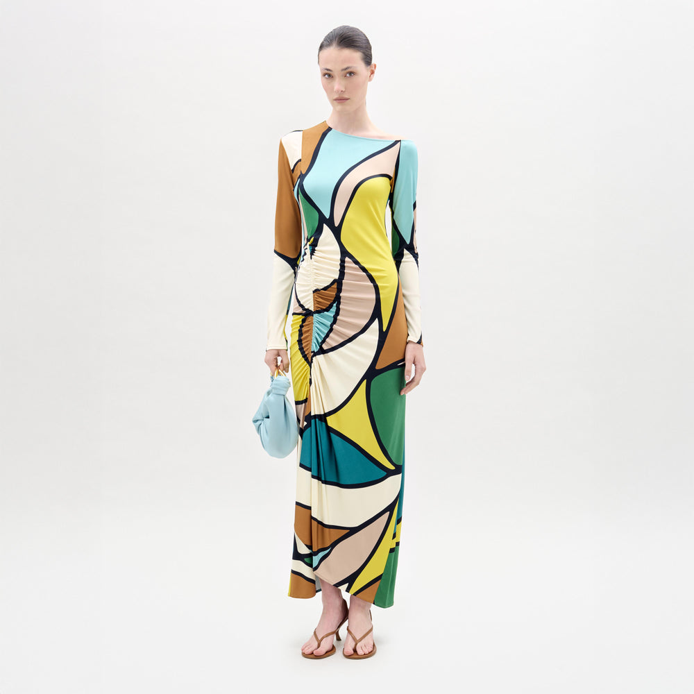 A woman stands against a white background wearing the Ilania Dress, a long midi with an abstract pattern in yellow, green, blue, brown, and black Italian fabric. She holds a small blue bag and wears brown sandals.