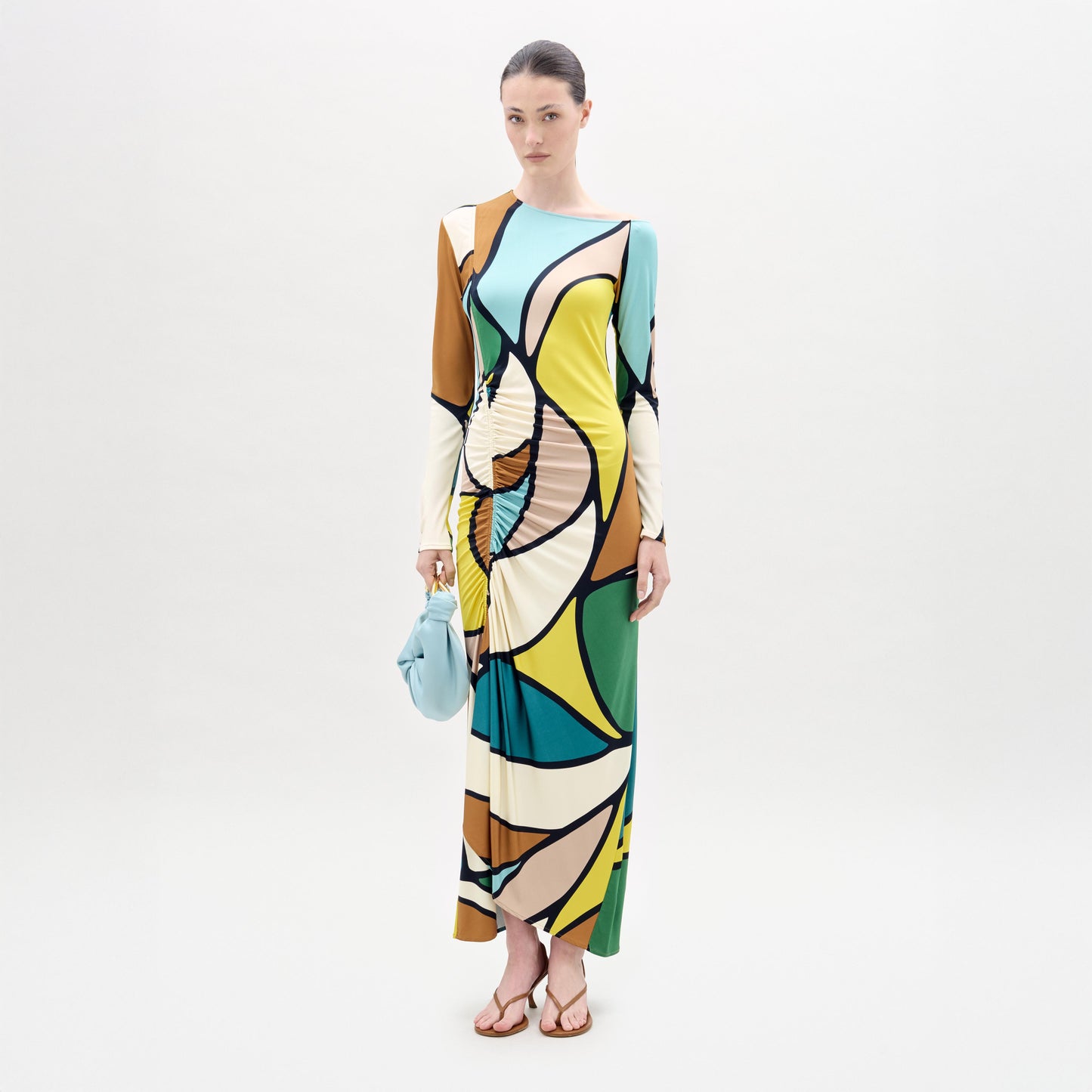 A woman stands against a white background wearing the Ilania Dress, a long midi with an abstract pattern in yellow, green, blue, brown, and black Italian fabric. She holds a small blue bag and wears brown sandals.