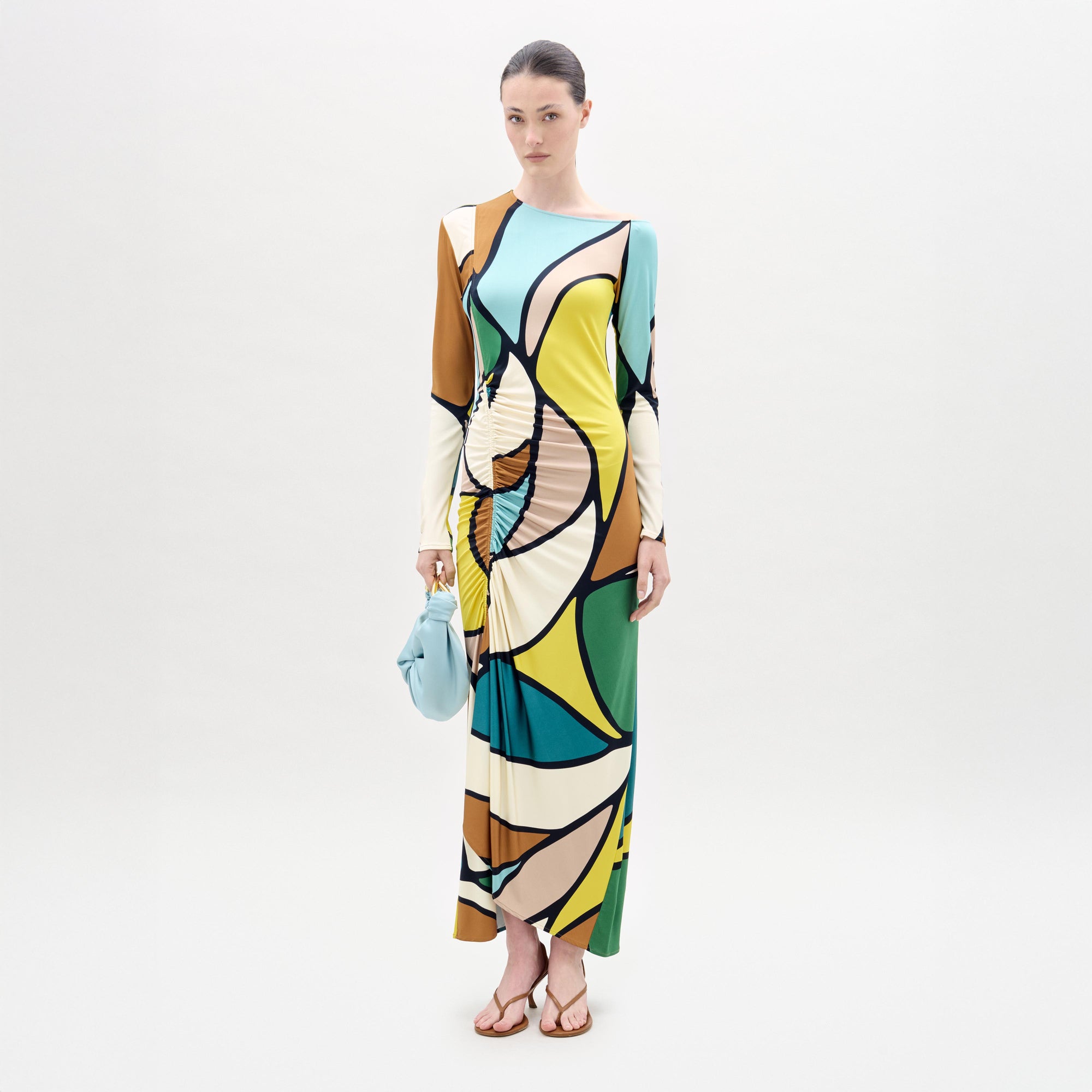 A woman stands against a white background wearing the Ilania Dress, a long midi with an abstract pattern in yellow, green, blue, brown, and black Italian fabric. She holds a small blue bag and wears brown sandals.