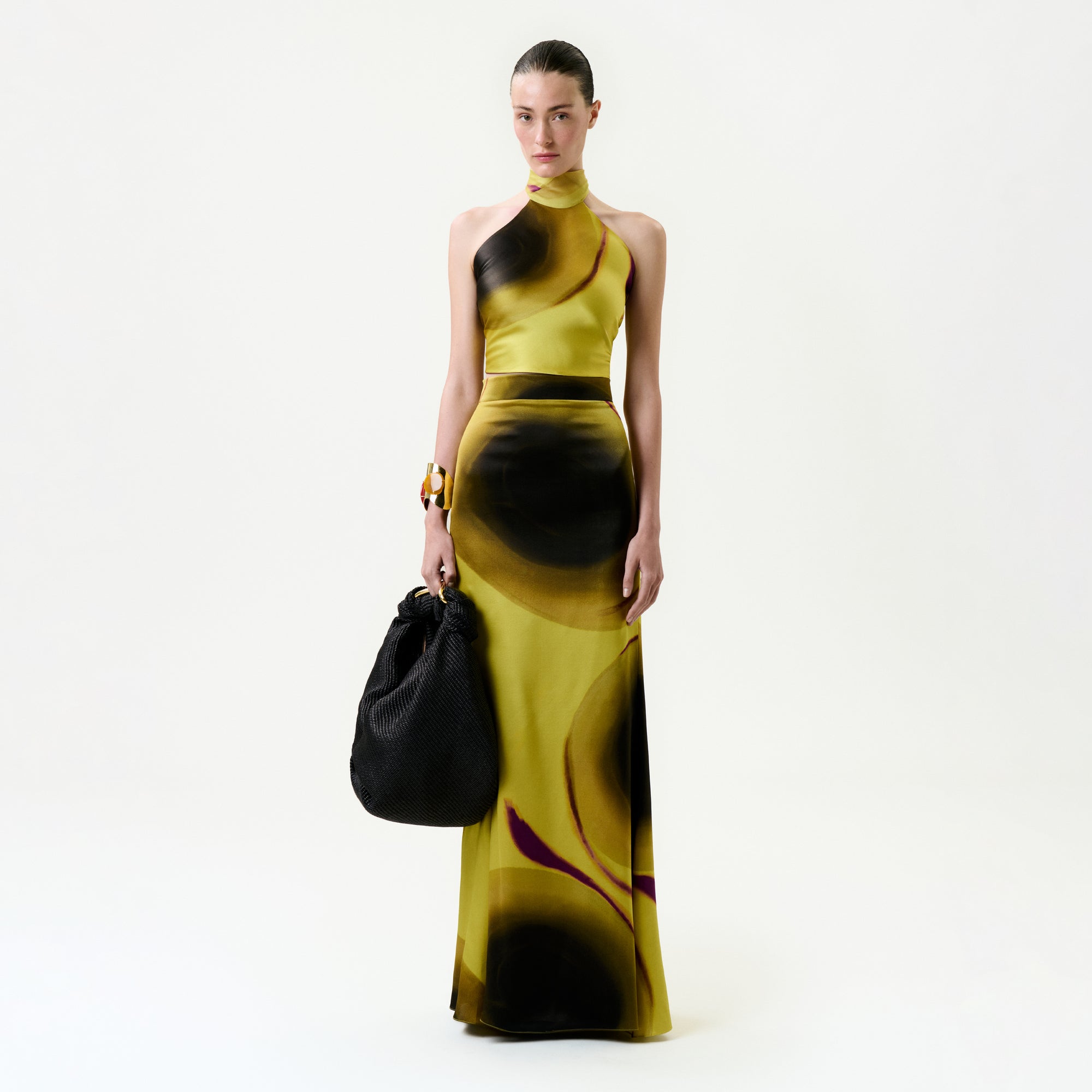 A woman stands against a plain white background wearing the Marisol Skirt, a yellow floor-length silk skirt with black and purple abstract patterns, holding a large textured black bag and accessorized with a chunky gold bracelet.