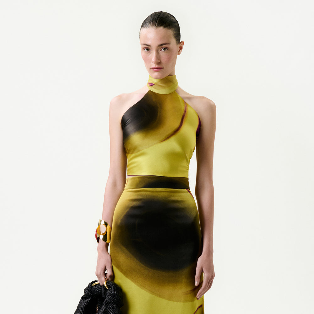 A woman with slicked-back hair wears the Nair Blouse in yellow Italian fabric with abstract brown and black patterns, accessorized with a chunky bracelet and a textured black bag, standing against a plain light backdrop.