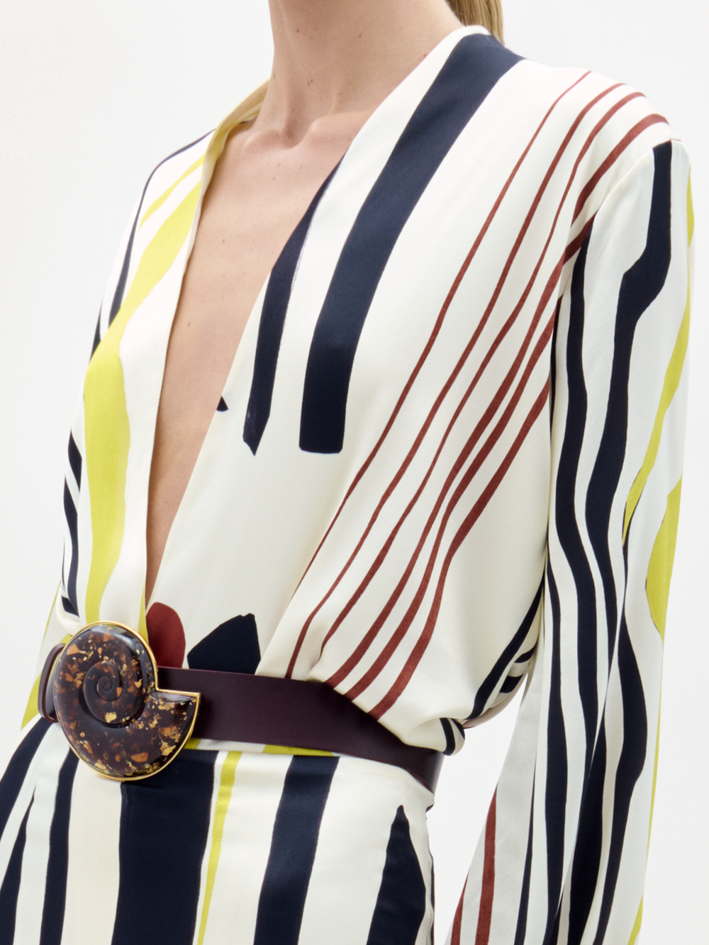A woman poses against a plain background wearing the Honey Blouse, featuring long sleeves and a deep V-neck with black, white, yellow, and red stripes, styled with a wide belt.