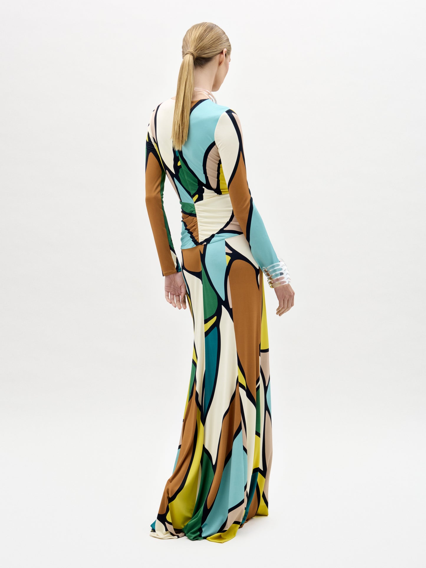 A woman models the Lucian Dress—a maxi with a form-fitting bodice, colorful abstract pattern, and front twist detail—against a plain white background.