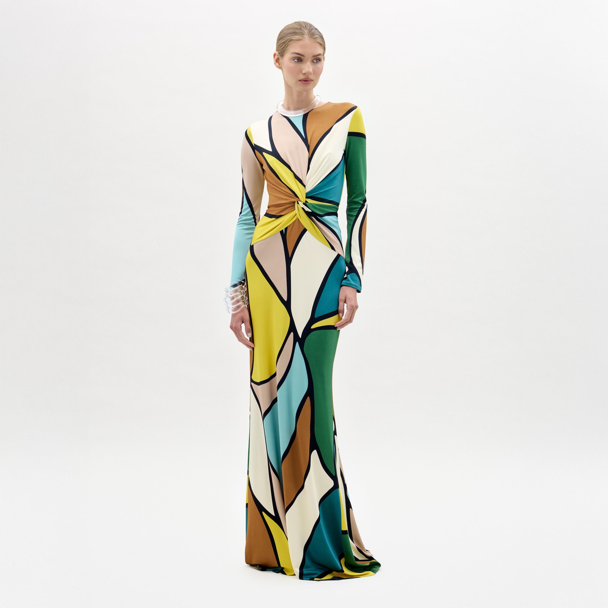 A woman models the Lucian Dress—a maxi with a form-fitting bodice, colorful abstract pattern, and front twist detail—against a plain white background.