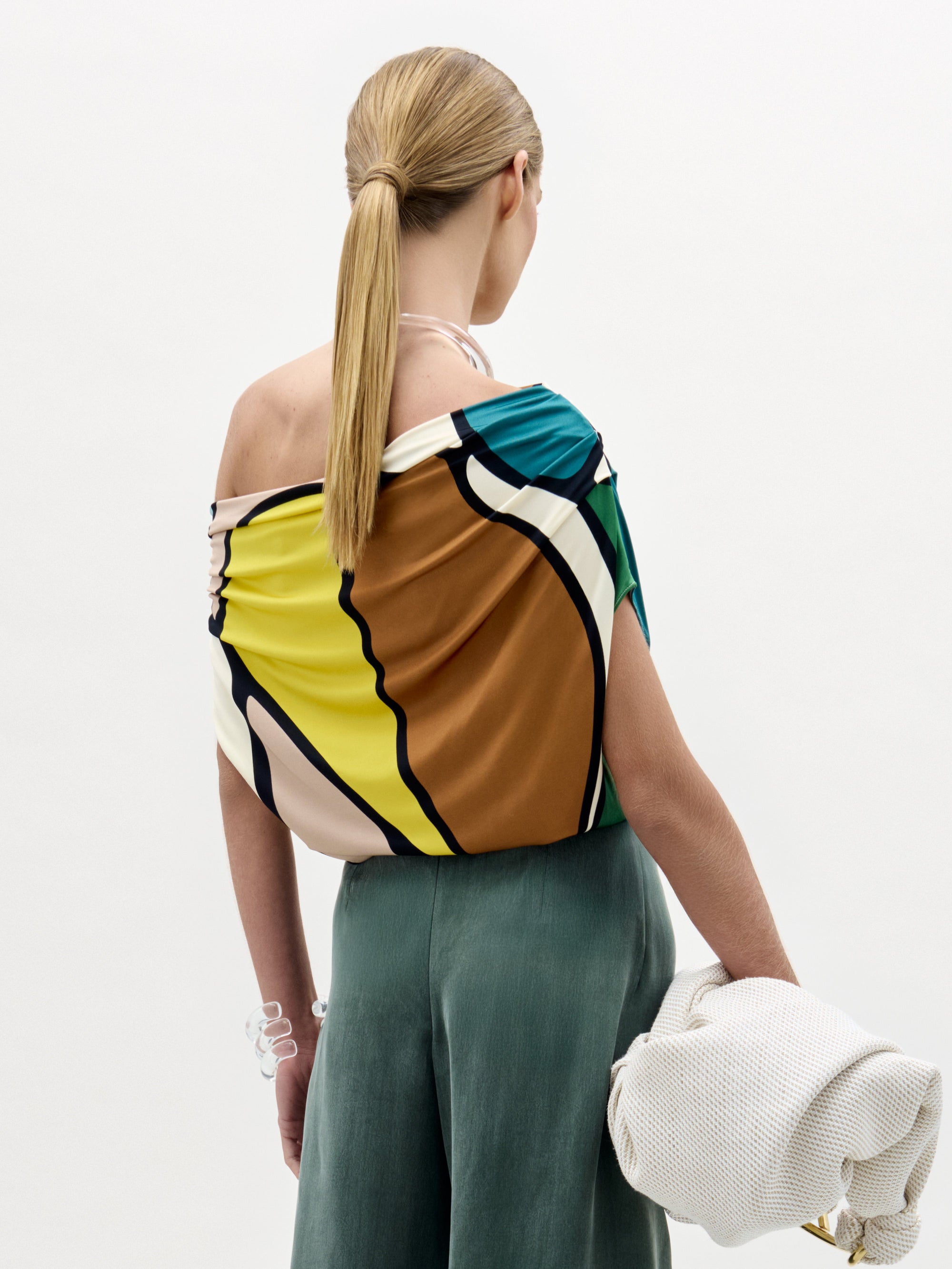 A woman stands against a plain background wearing the colorful Indiana Blouse with an off-the-shoulder neckline, a green high-waisted skirt with a bow, and holding a white textured bag.