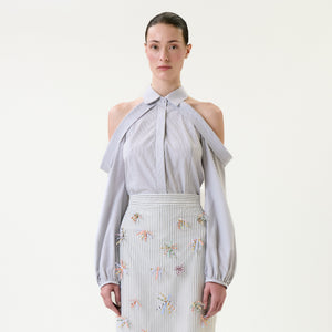 A woman wears the Nurith Shirt, a light blue cold-shoulder blouse with draped sleeves, paired with a matching high-waisted skirt adorned with small, colorful embroidered flowers, standing against a plain white background.