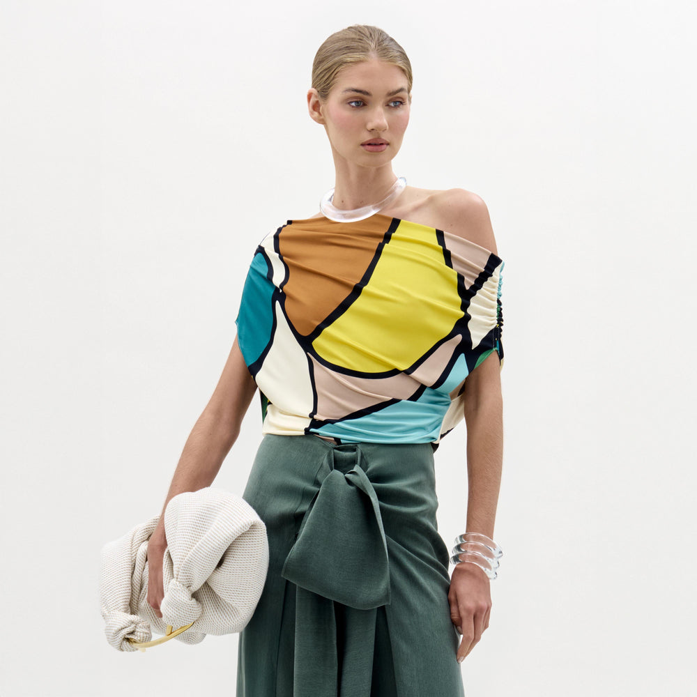 A woman stands against a plain background wearing the colorful Indiana Blouse with an off-the-shoulder neckline, a green high-waisted skirt with a bow, and holding a white textured bag.