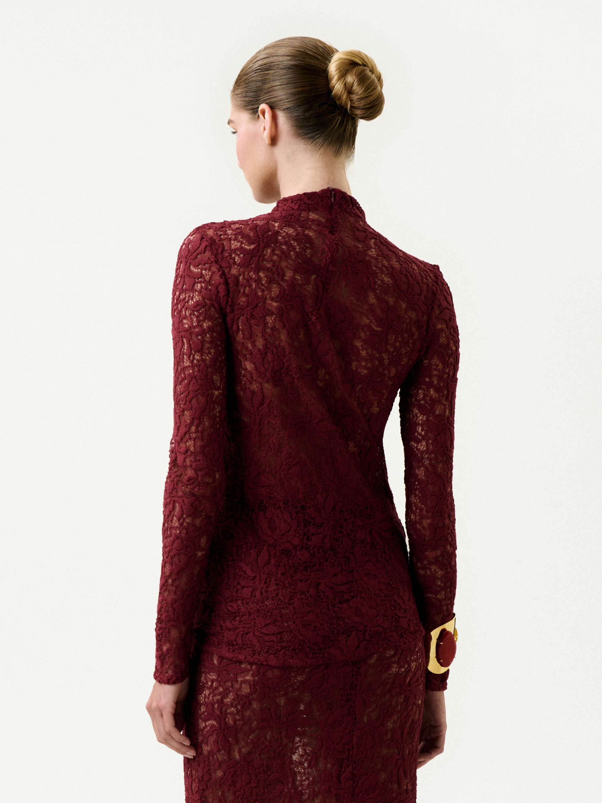 A woman with her hair in a neat bun, wearing the Lisibel Blouse—an elegant, long-sleeved burgundy lace top made from Italian fabric—stands facing away from the camera against a plain white background.