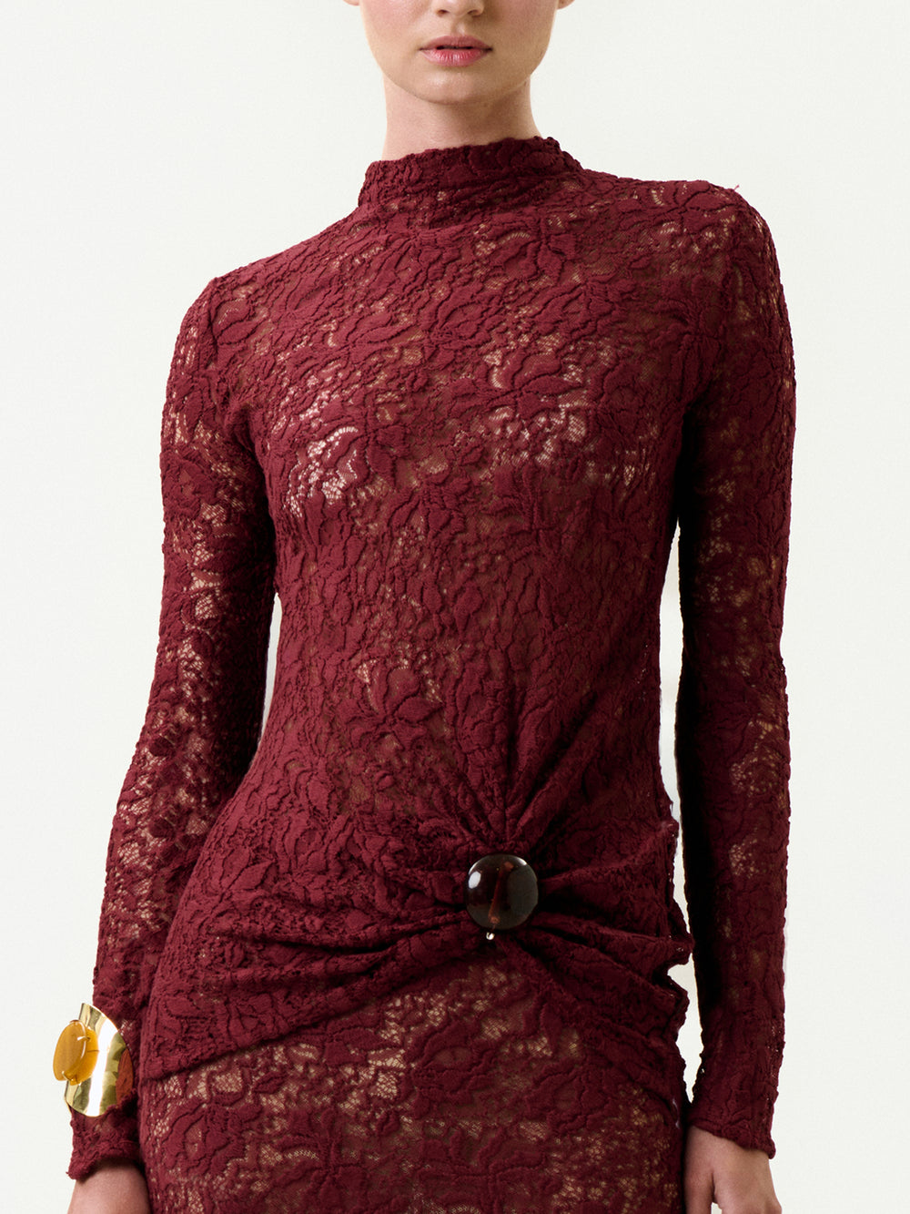 A person wearing the Lisibel Blouse—long-sleeved, burgundy lace, made from luxurious Italian fabric, with a high neckline, gathered waist, and a large round button—paired with a chunky gold bracelet.