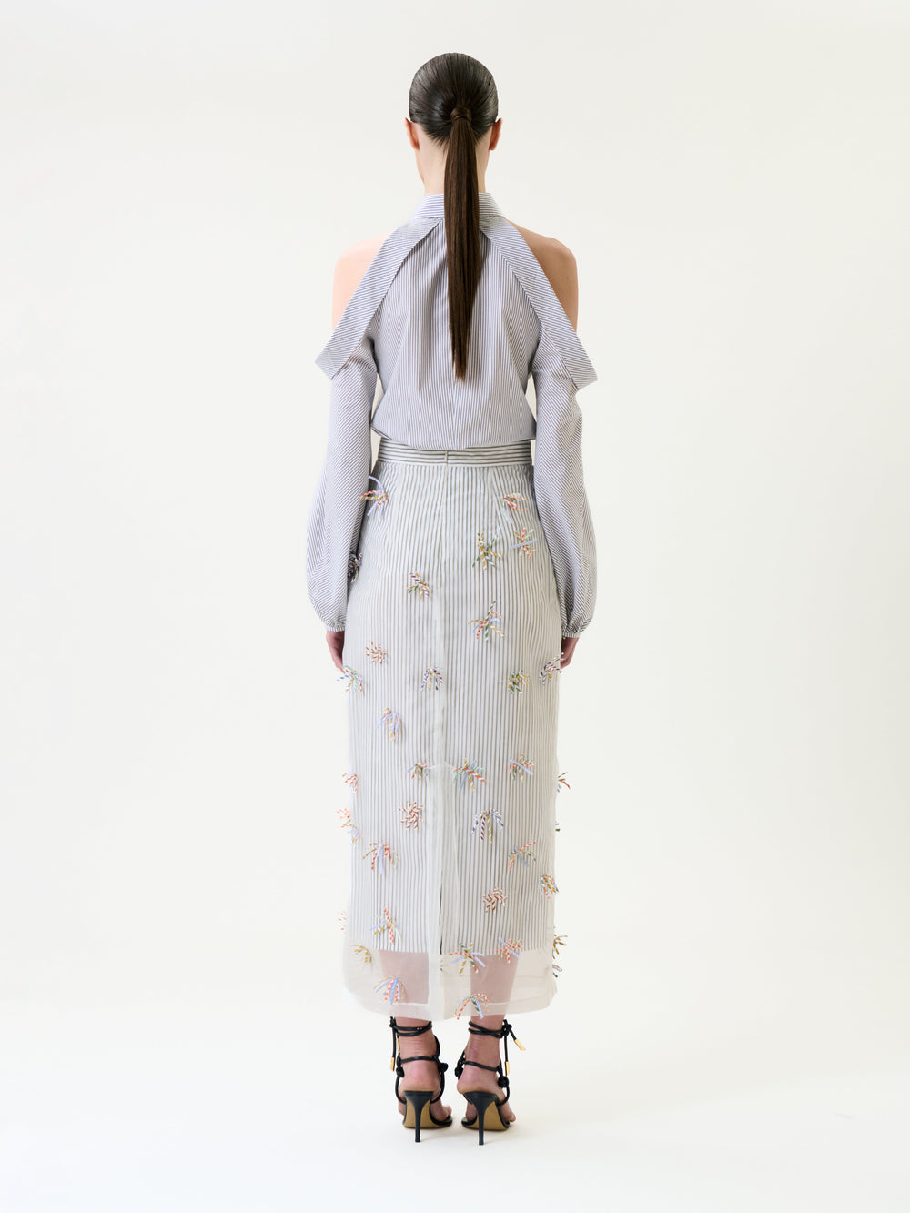 A woman with long, straight hair stands facing away, wearing a light gray, long-sleeve, open-shoulder blouse and the Uma Skirt—a sheer, embroidered midi skirt with floral details—paired with black strappy heeled sandals.