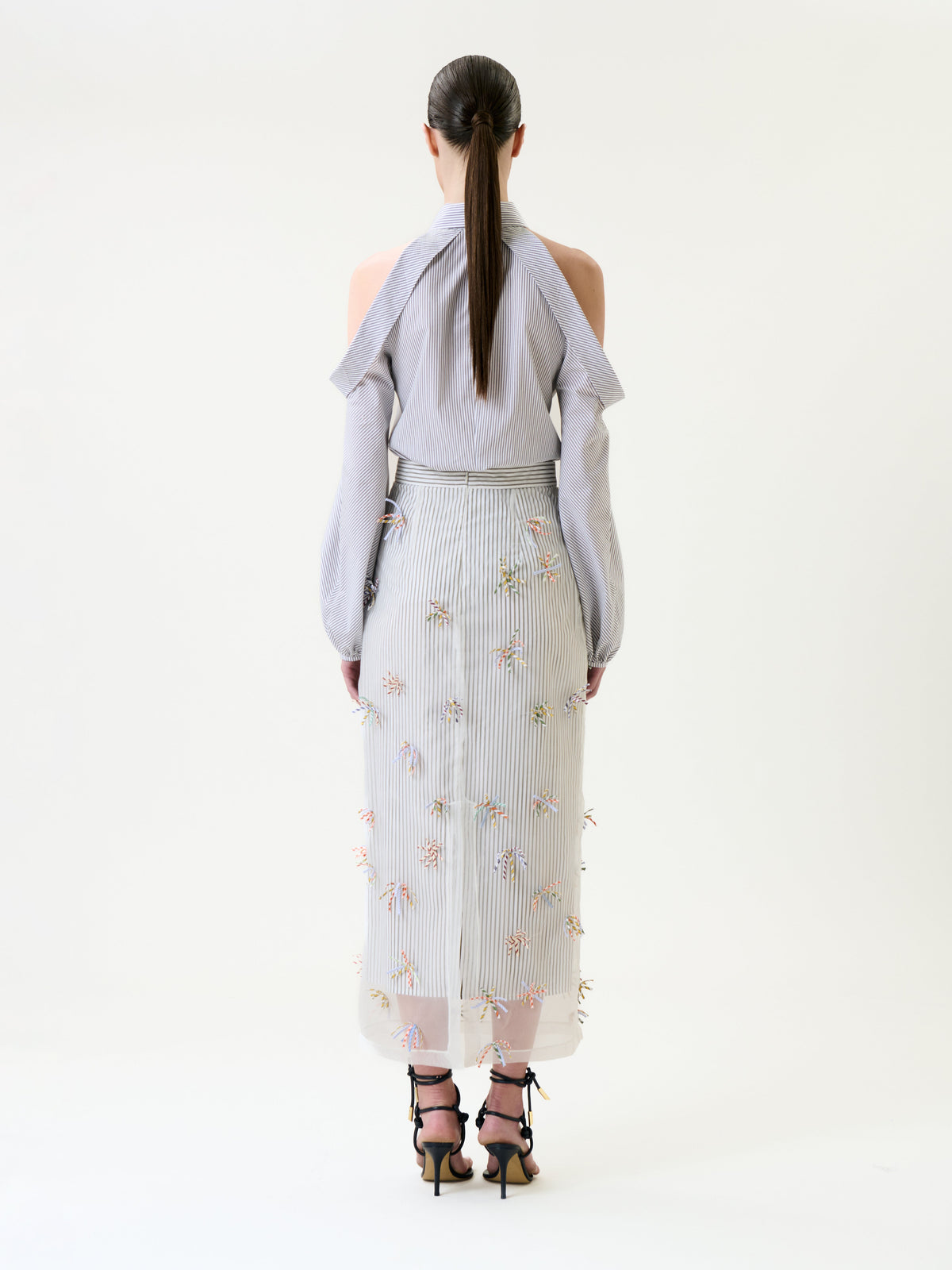 A woman with long, straight hair stands facing away, wearing a light gray, long-sleeve, open-shoulder blouse and the Uma Skirt—a sheer, embroidered midi skirt with floral details—paired with black strappy heeled sandals.