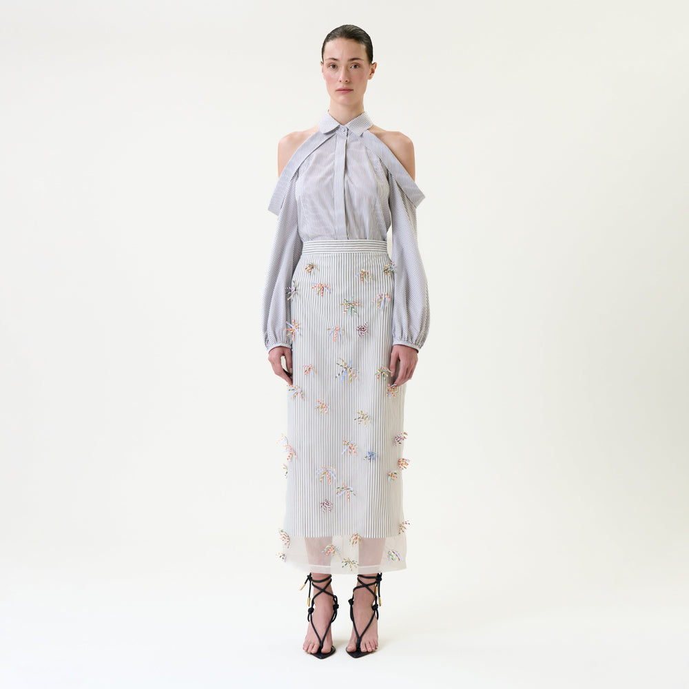 A woman stands against a plain background wearing a light blue, cold-shoulder blouse and the Uma Skirt—a sheer embroidered skirt with floral patterns—paired with black strappy sandals.