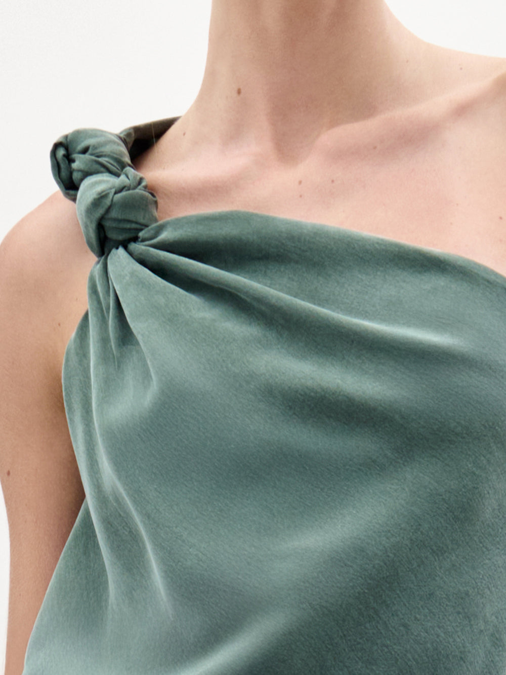 A woman stands against a white background wearing the Yanet Blouse, a sage green, one-shoulder top with an asymmetric neckline, knot detail at the shoulder, Italian fabric, and a large bow accent at the waist.