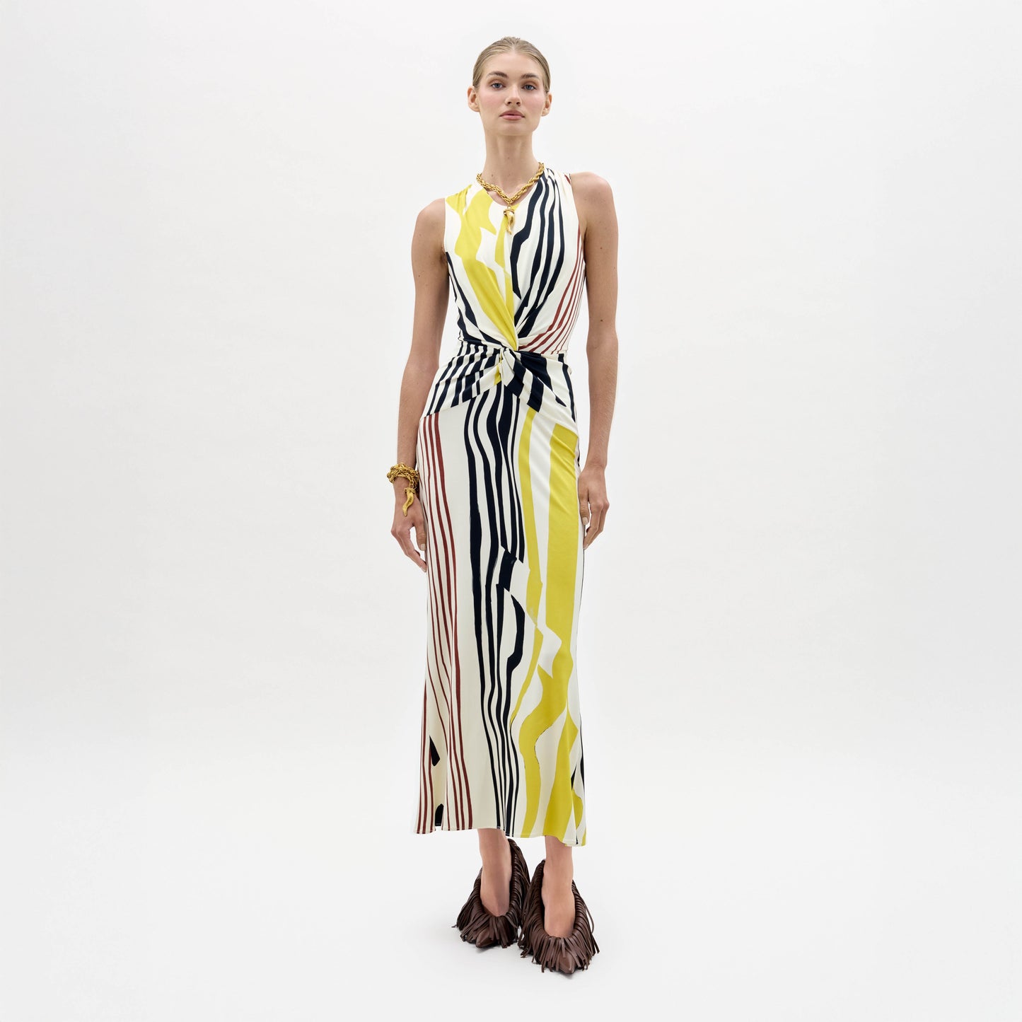 A woman stands against a white background wearing the Luna Dress—a sleeveless, ankle-length dress with yellow, black, and white vertical abstract stripes—and brown fringed shoes.