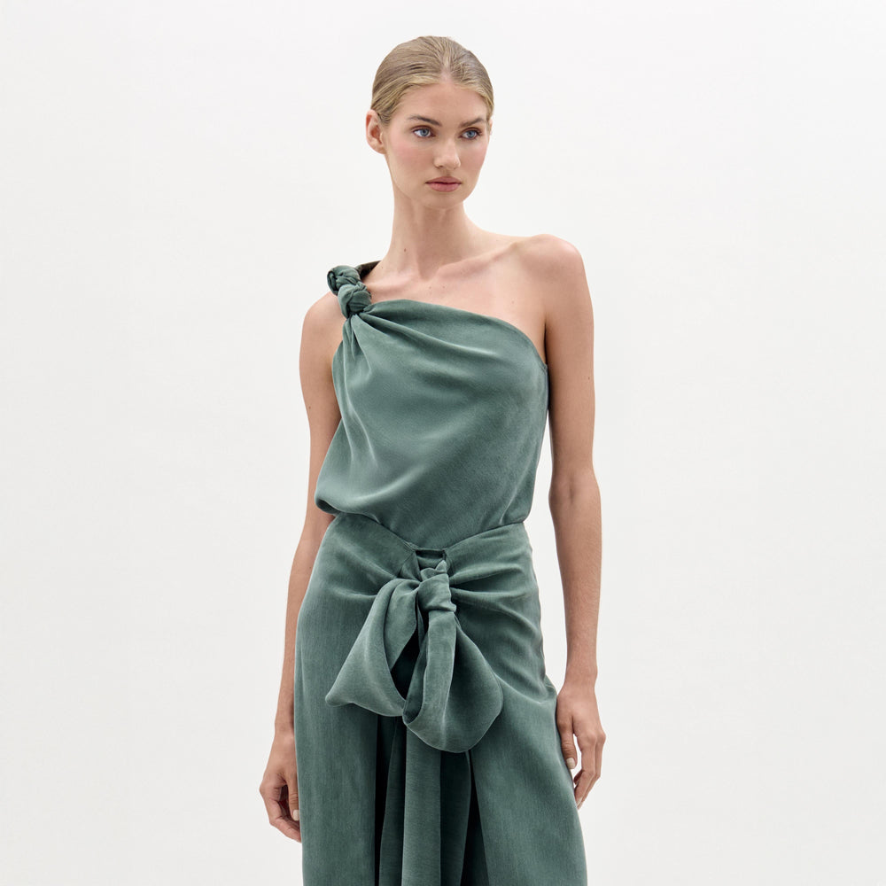 A woman stands against a white background wearing the Yanet Blouse, a sage green, one-shoulder top with an asymmetric neckline, knot detail at the shoulder, Italian fabric, and a large bow accent at the waist.