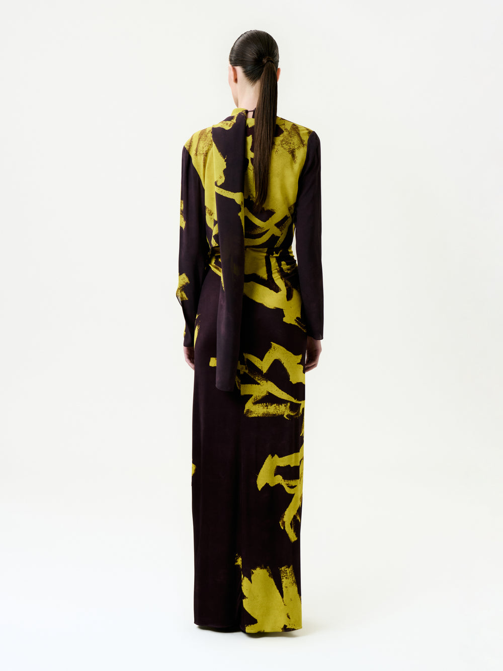 A person with long hair in a low ponytail faces away, wearing the floor-length, long-sleeved Luisanna Cape adorned with bold yellow abstract patterns on luxurious Italian fabric, standing in a plain white studio.