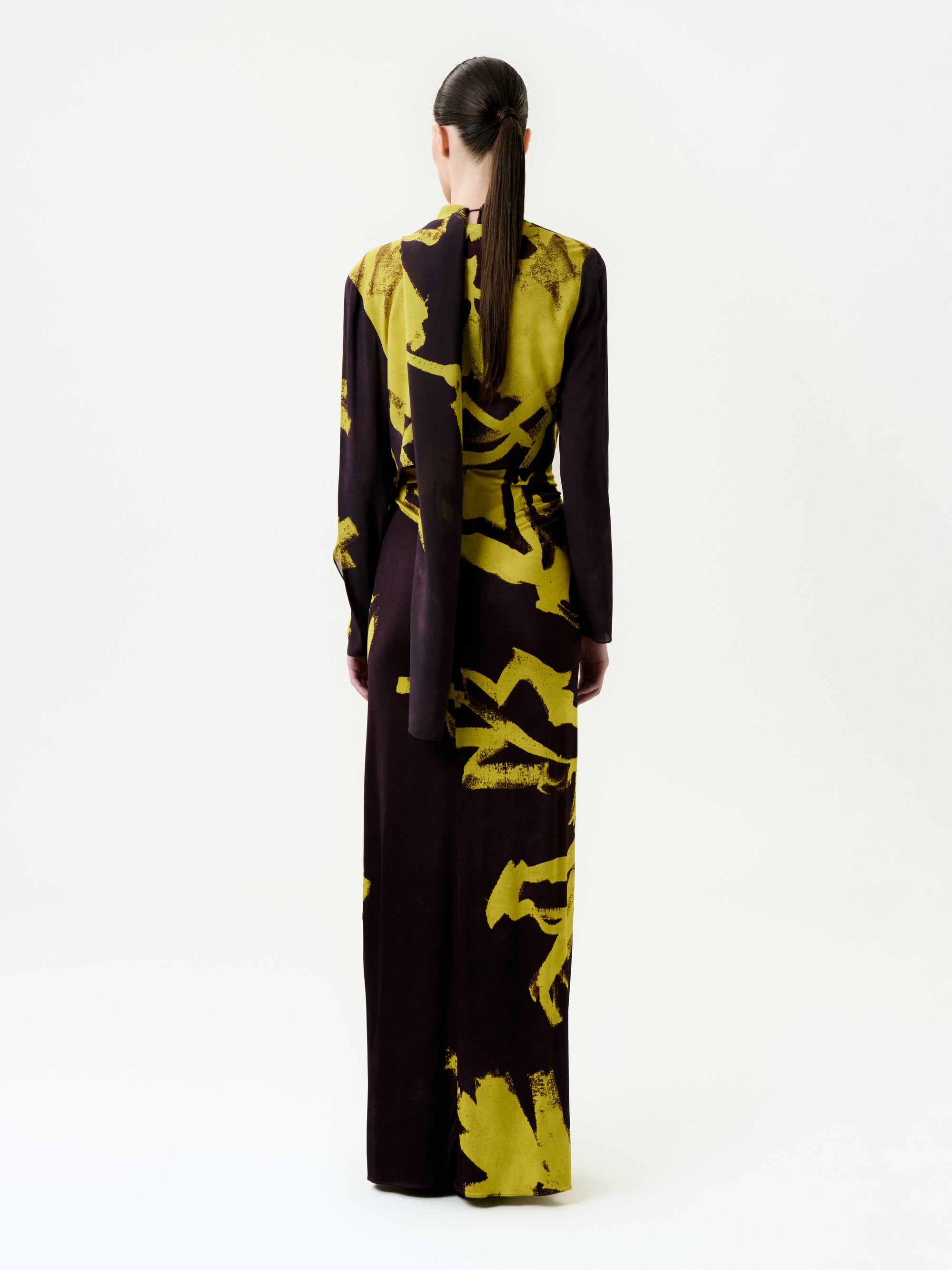 A person with long hair in a low ponytail faces away, wearing the floor-length, long-sleeved Luisanna Cape adorned with bold yellow abstract patterns on luxurious Italian fabric, standing in a plain white studio.