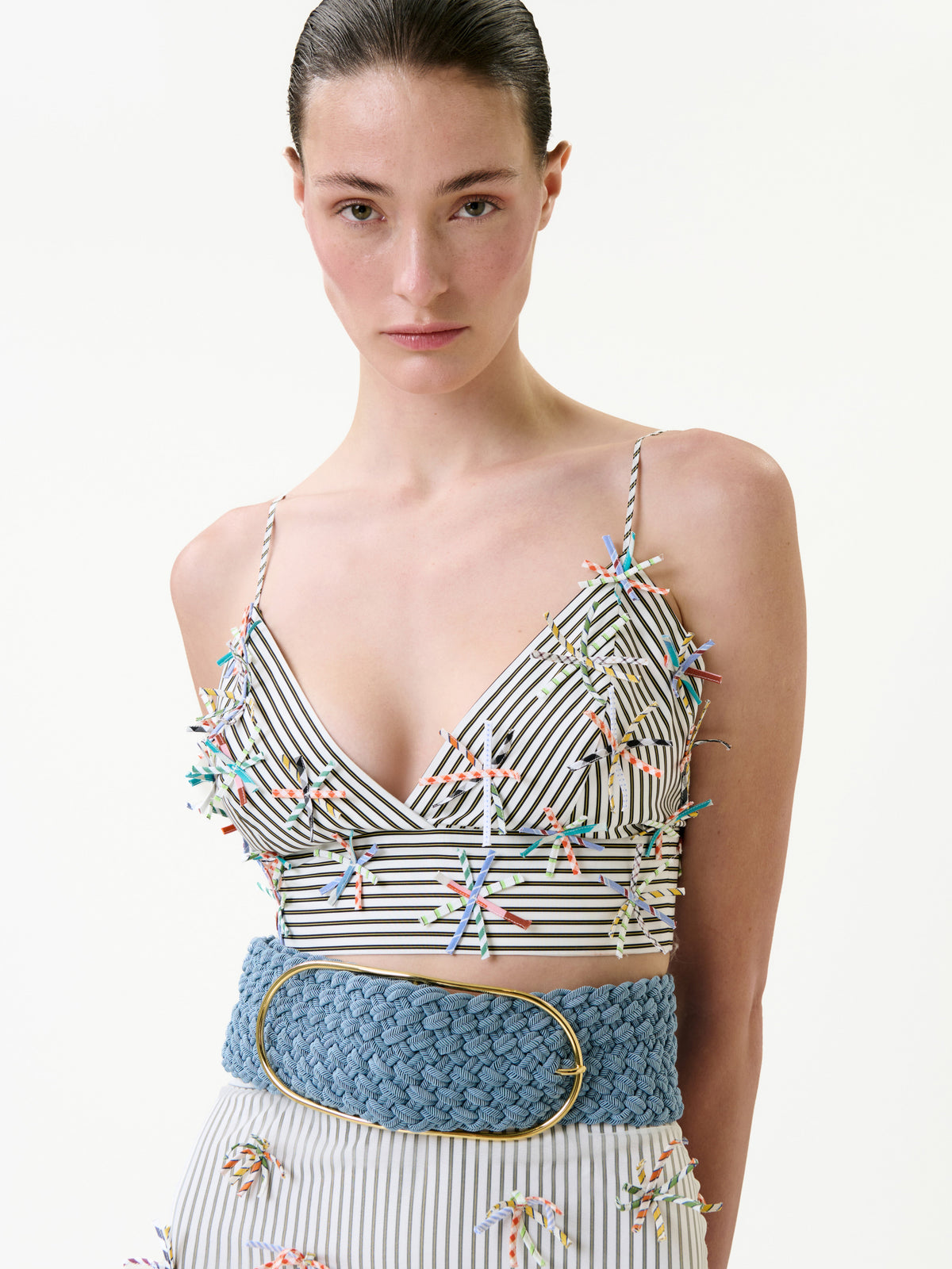 A woman wearing the Ibbie Top, featuring black-and-white stripes with multicolored embellishments and a wide blue braided belt, stands against a plain white background.