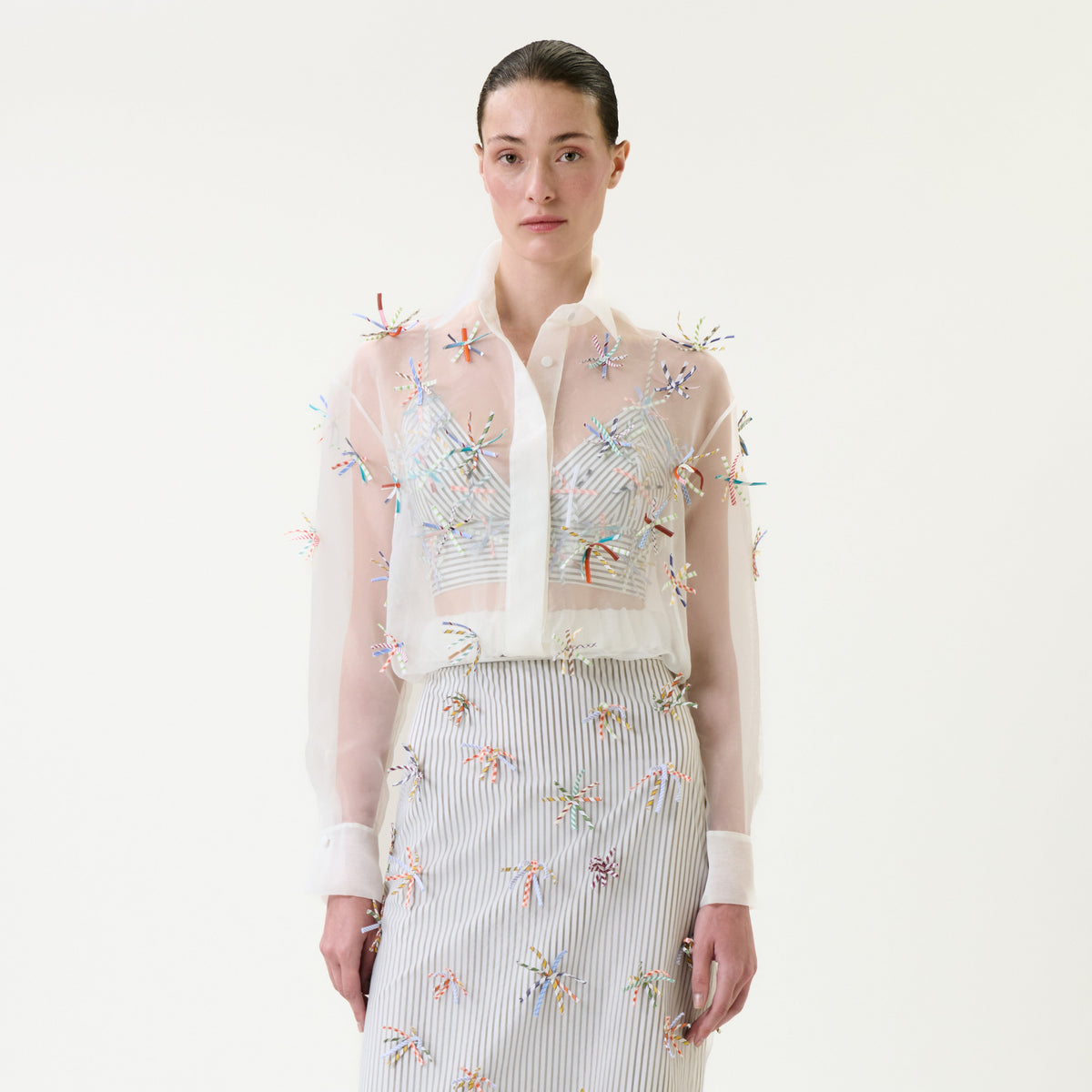 A woman wearing the Katniss Jacket—a sheer, white, long-sleeve blouse with colorful embroidered details—paired with a matching striped skirt, standing against a plain light background.