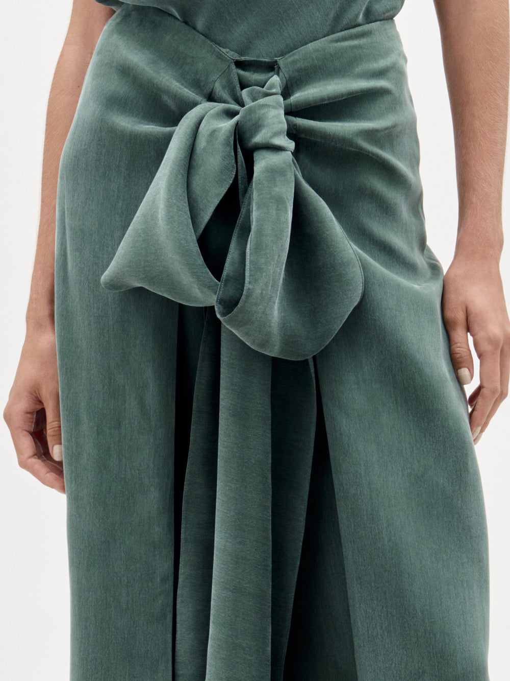 A woman stands against a plain light background wearing the Arenzano Pant, a sage green, one-shoulder jumpsuit made from Italian fabric with a high-rise waist, wide legs, and a statement bow at the waist.