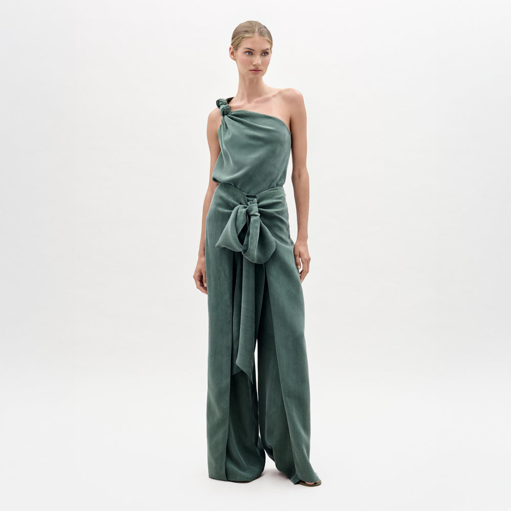 A woman stands against a plain light background wearing the Arenzano Pant, a sage green, one-shoulder jumpsuit made from Italian fabric with a high-rise waist, wide legs, and a statement bow at the waist.
