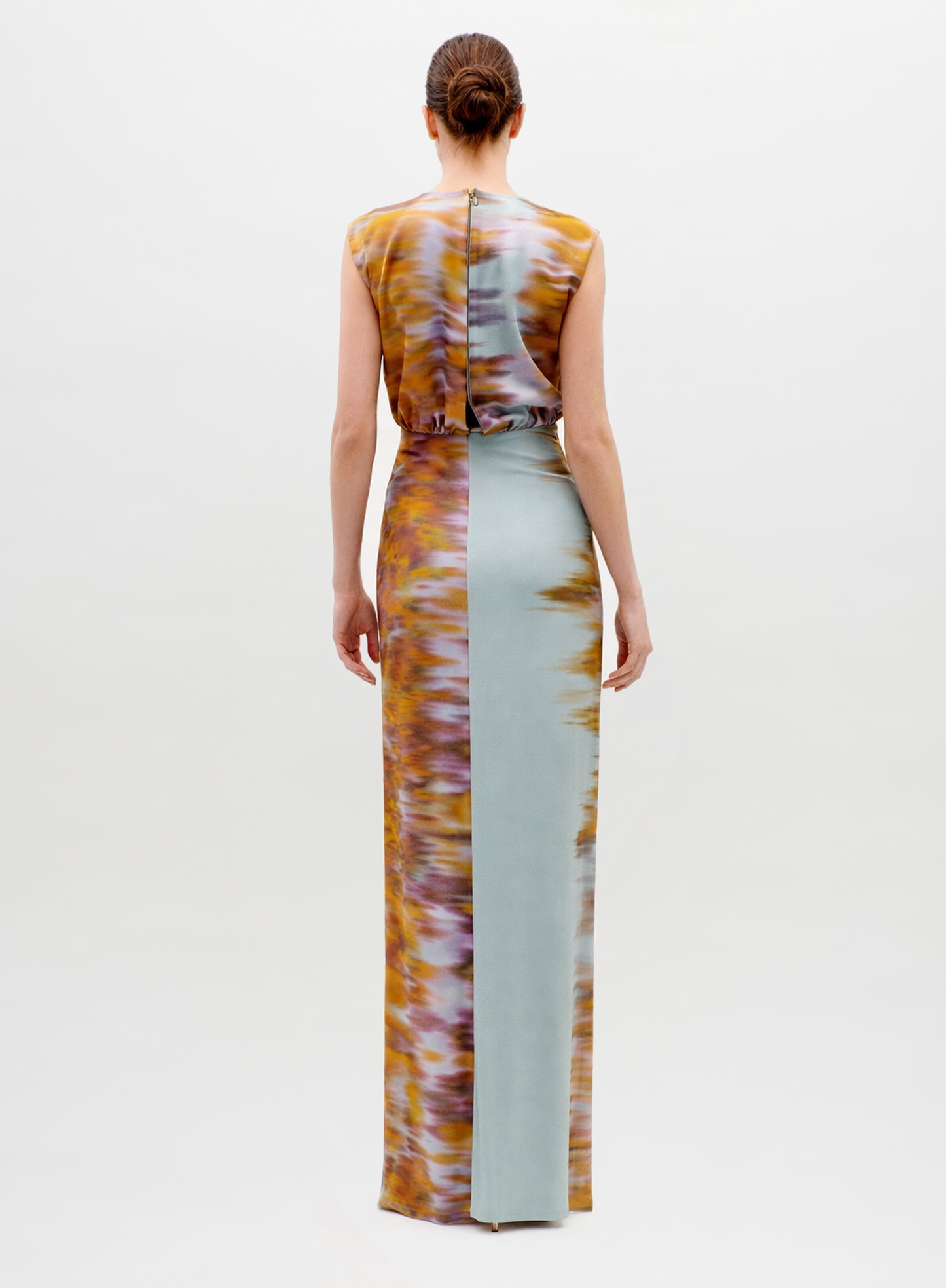 A woman with a neat low bun faces away, wearing the sleeveless, floor-length Winona Dress featuring a split design: one side in light blue and the other in a brown and yellow textured pattern.