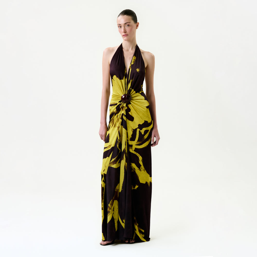 A woman stands against a white background wearing the Aiko Dress, a bold yellow and black floral maxi with a central waist knot, crafted from premium Italian fabric. Her hair is pulled back as she looks straight ahead.