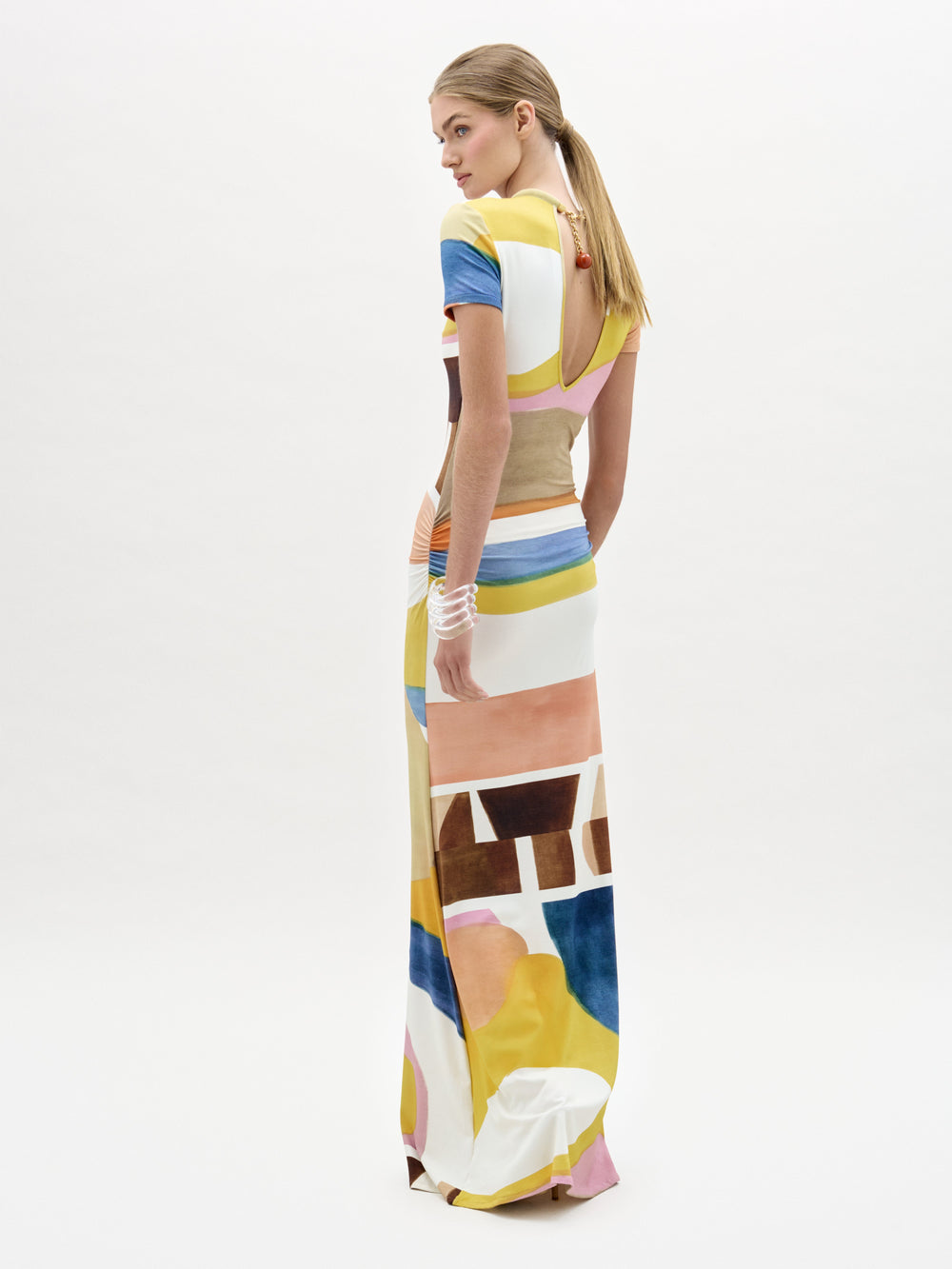 A woman stands against a white background wearing the Ikia Dress, a floor-length, short-sleeved maxi crafted from Italian fabric in a colorful abstract pattern of yellow, pink, blue, brown, and white shapes.