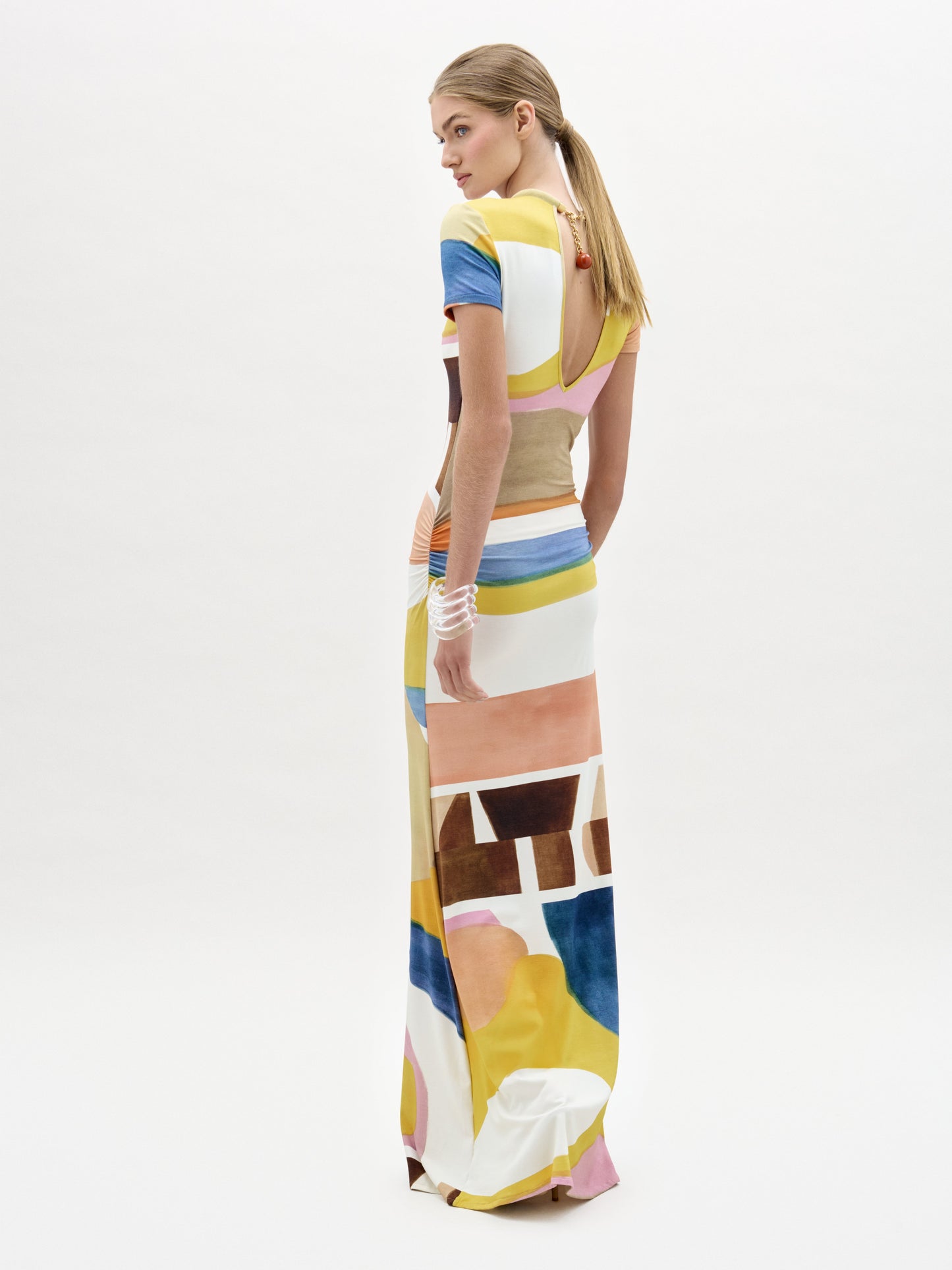 A woman stands against a white background wearing the Ikia Dress, a floor-length, short-sleeved maxi crafted from Italian fabric in a colorful abstract pattern of yellow, pink, blue, brown, and white shapes.