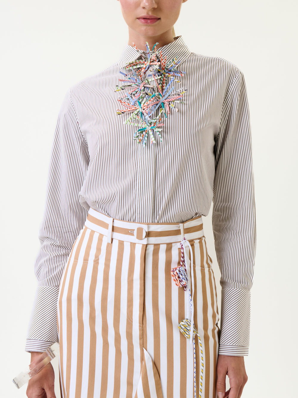 A person wearing the Karena Shirt—a white button-up shirt with thin black stripes—paired with tan-and-white striped high-waisted pants. Colorful decorative pieces hang from the collar and waistband. The background is plain white.