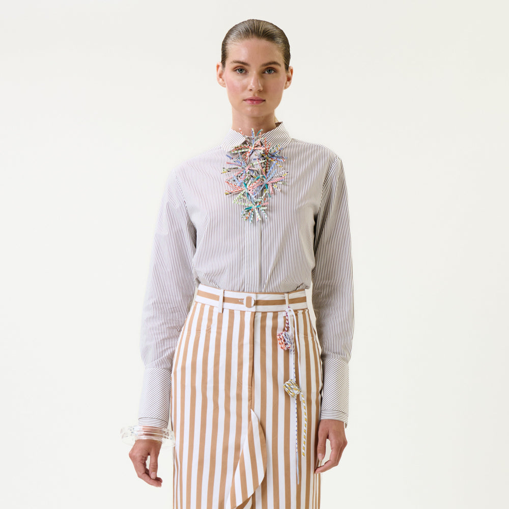A person stands facing forward wearing the Karena Shirt, which features colorful decorative embellishments and stripes, paired with a tan-and-white vertically striped skirt against a plain light background.