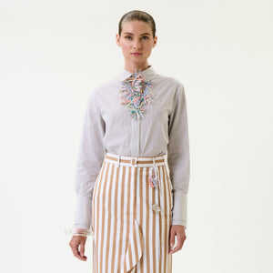 A person stands facing forward wearing the Karena Shirt, which features colorful decorative embellishments and stripes, paired with a tan-and-white vertically striped skirt against a plain light background.