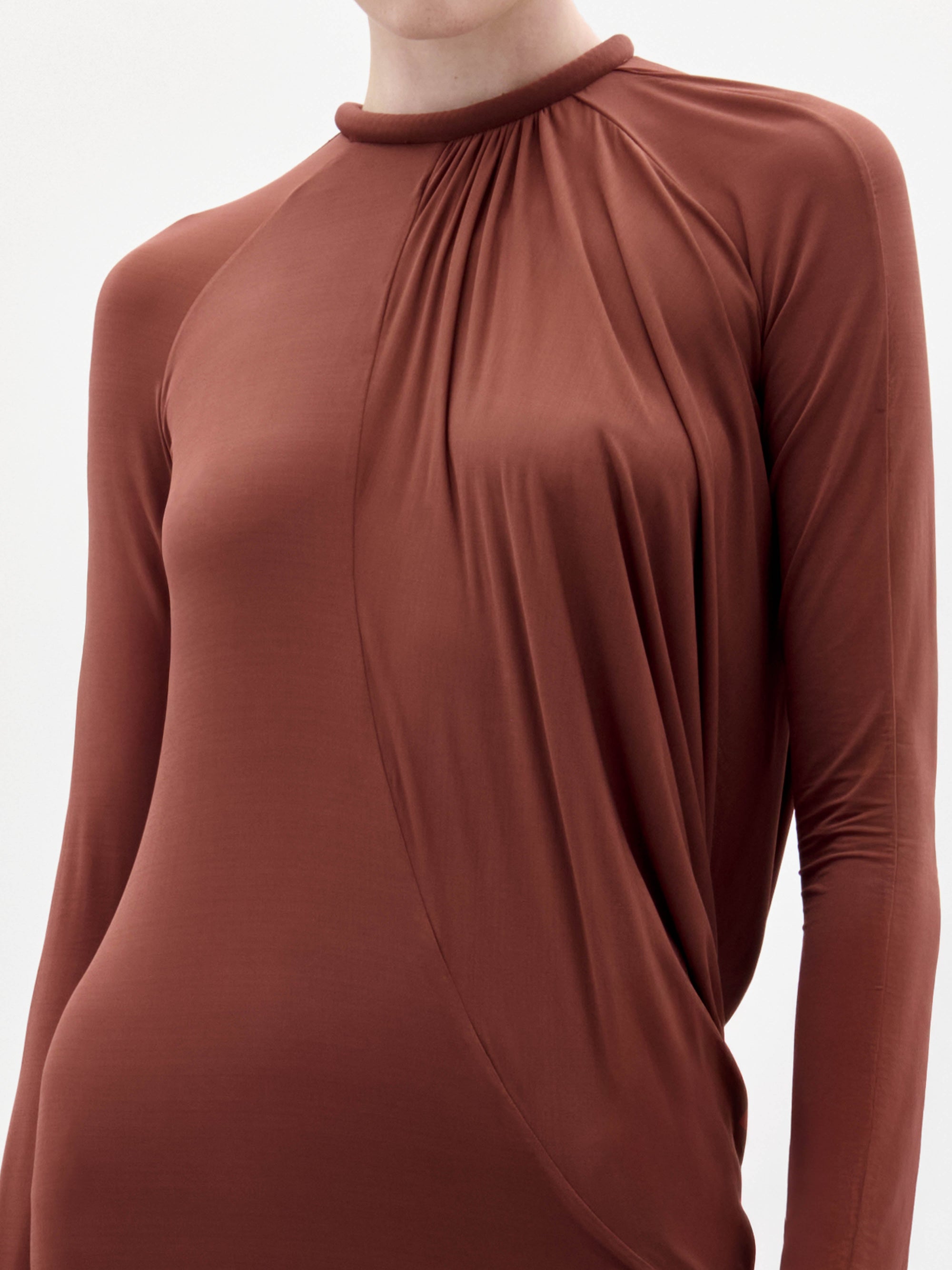 Someone stands against a plain white background wearing the Lais Dress, a long brown dress with long sleeves and a gathered neckline, styled with minimal accessories.