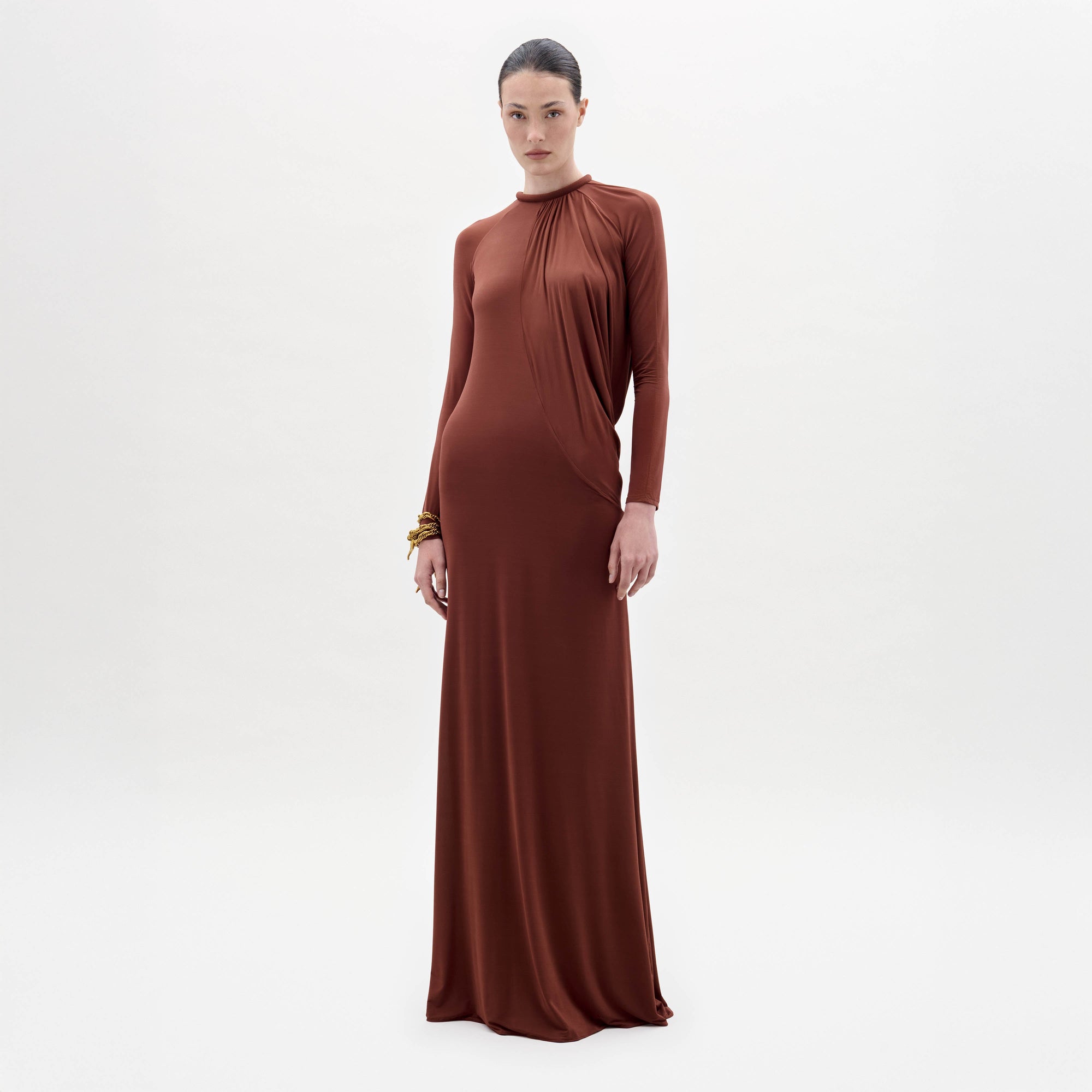 Someone stands against a plain white background wearing the Lais Dress, a long brown dress with long sleeves and a gathered neckline, styled with minimal accessories.