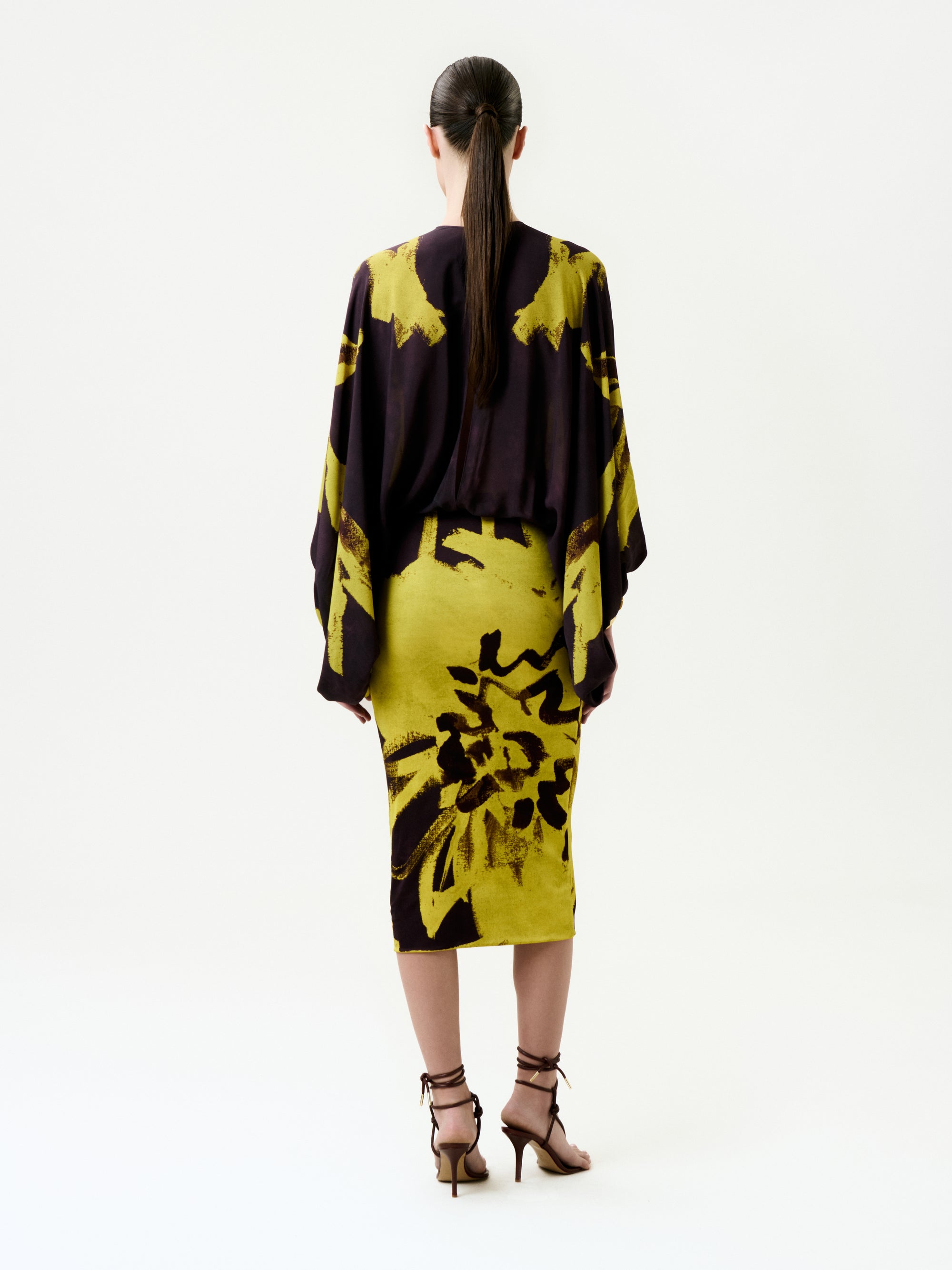 A woman with long dark hair in a ponytail is seen from behind wearing the Kumiko Dress, a knee-length piece with batwing sleeves, crafted from luxurious Italian fabric featuring bold yellow and dark purple abstract florals.