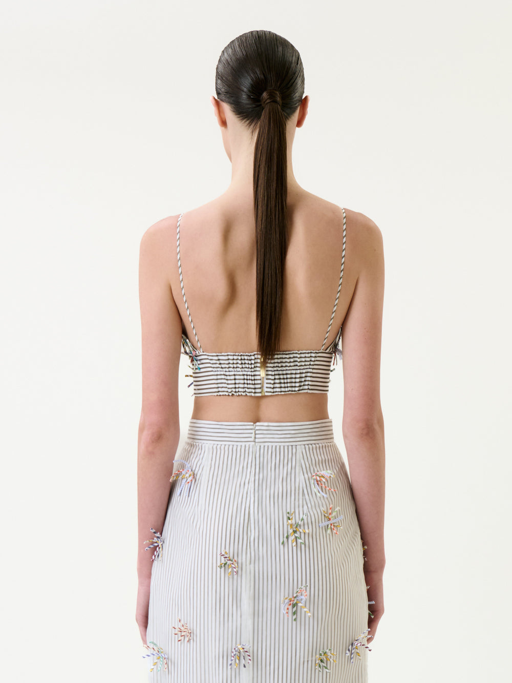 A woman with straight brown hair in a low ponytail is seen from behind against a plain white background, wearing the Ibbie Top—a striped crop top with thin straps—paired with a matching high-waisted skirt featuring small floral details.