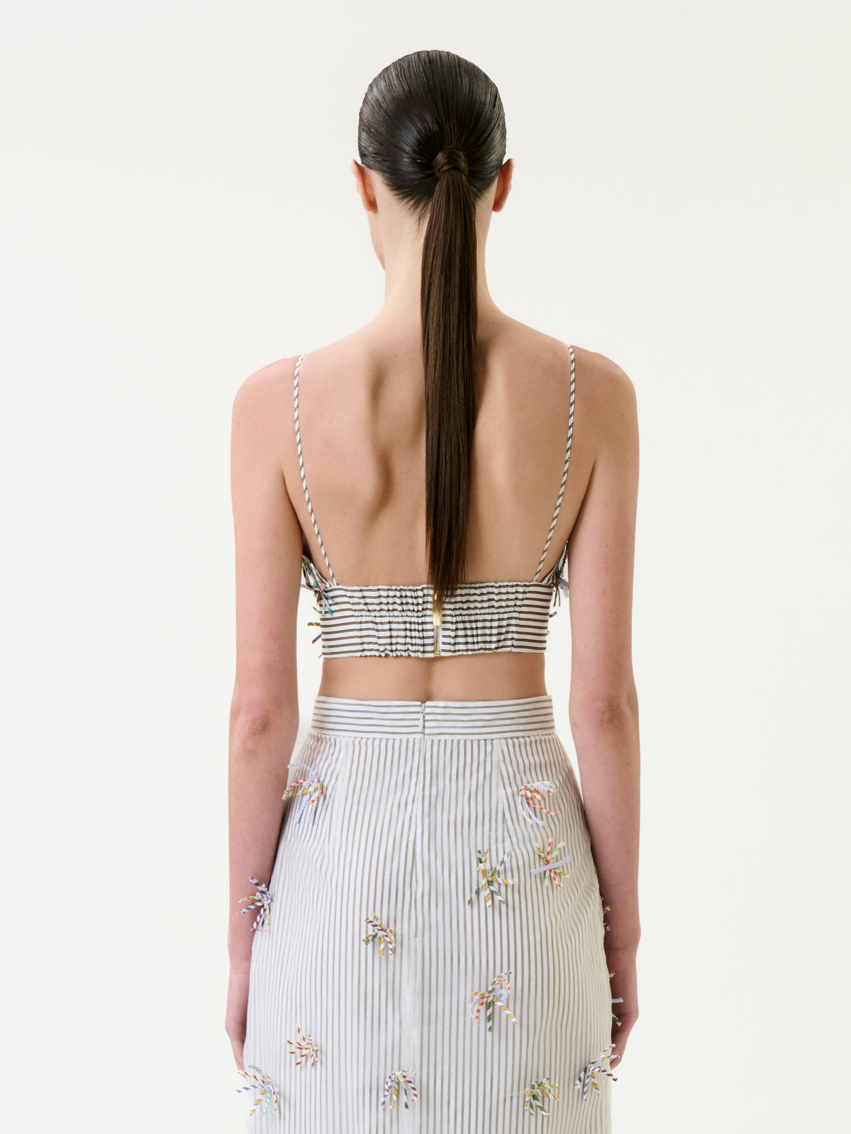 A woman with straight brown hair in a low ponytail is seen from behind against a plain white background, wearing the Ibbie Top—a striped crop top with thin straps—paired with a matching high-waisted skirt featuring small floral details.