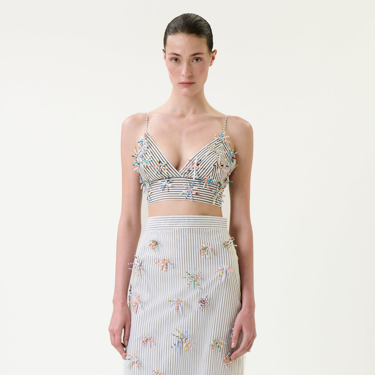 Wearing the Ibbie Top, a woman stands against a plain white background. The striped crop top is adorned with colorful 3D embellishments and paired with a matching skirt.