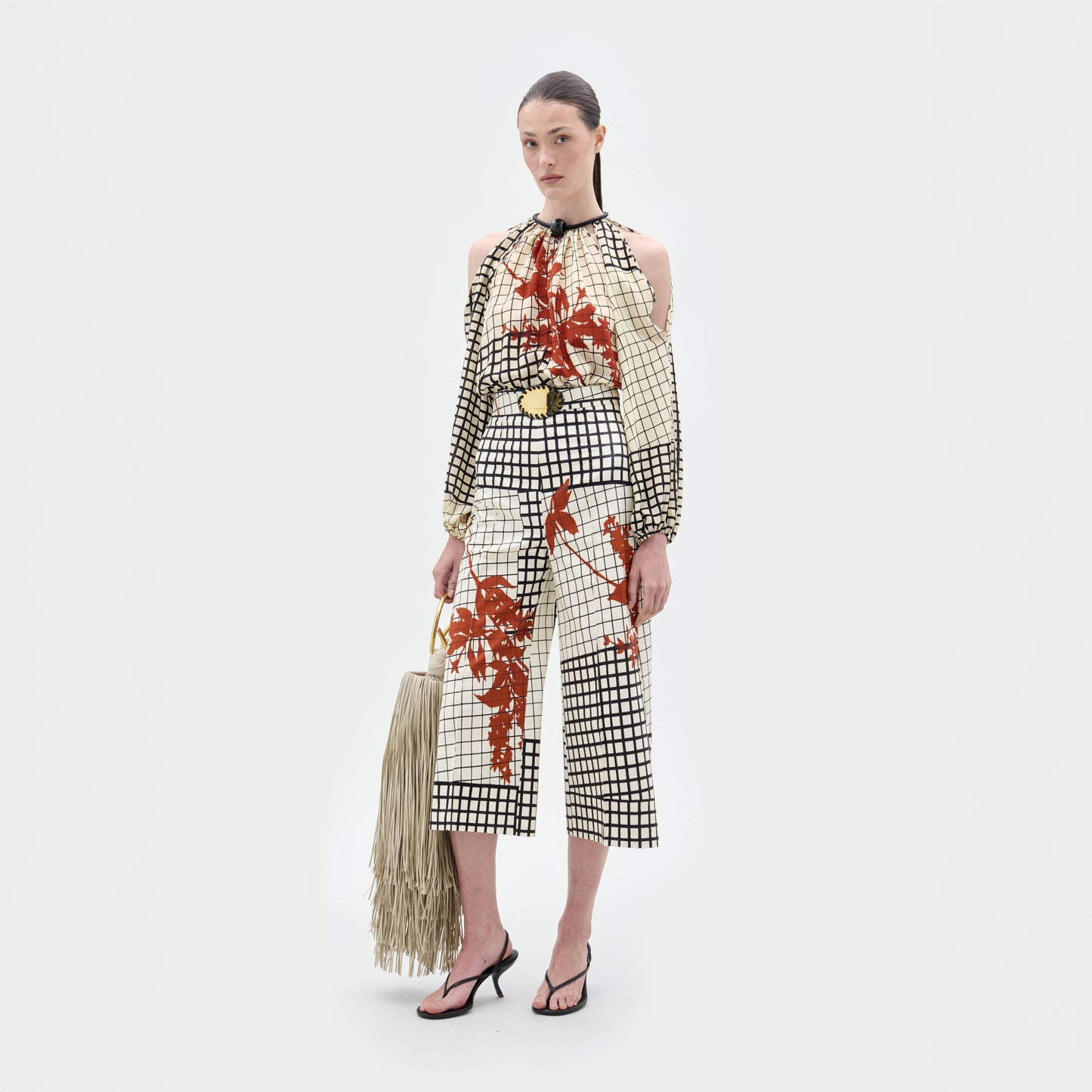 A woman stands against a plain background wearing the Ilma Pant, a grid-patterned outfit with red floral accents, paired with black sandals and holding a fringed cream handbag.