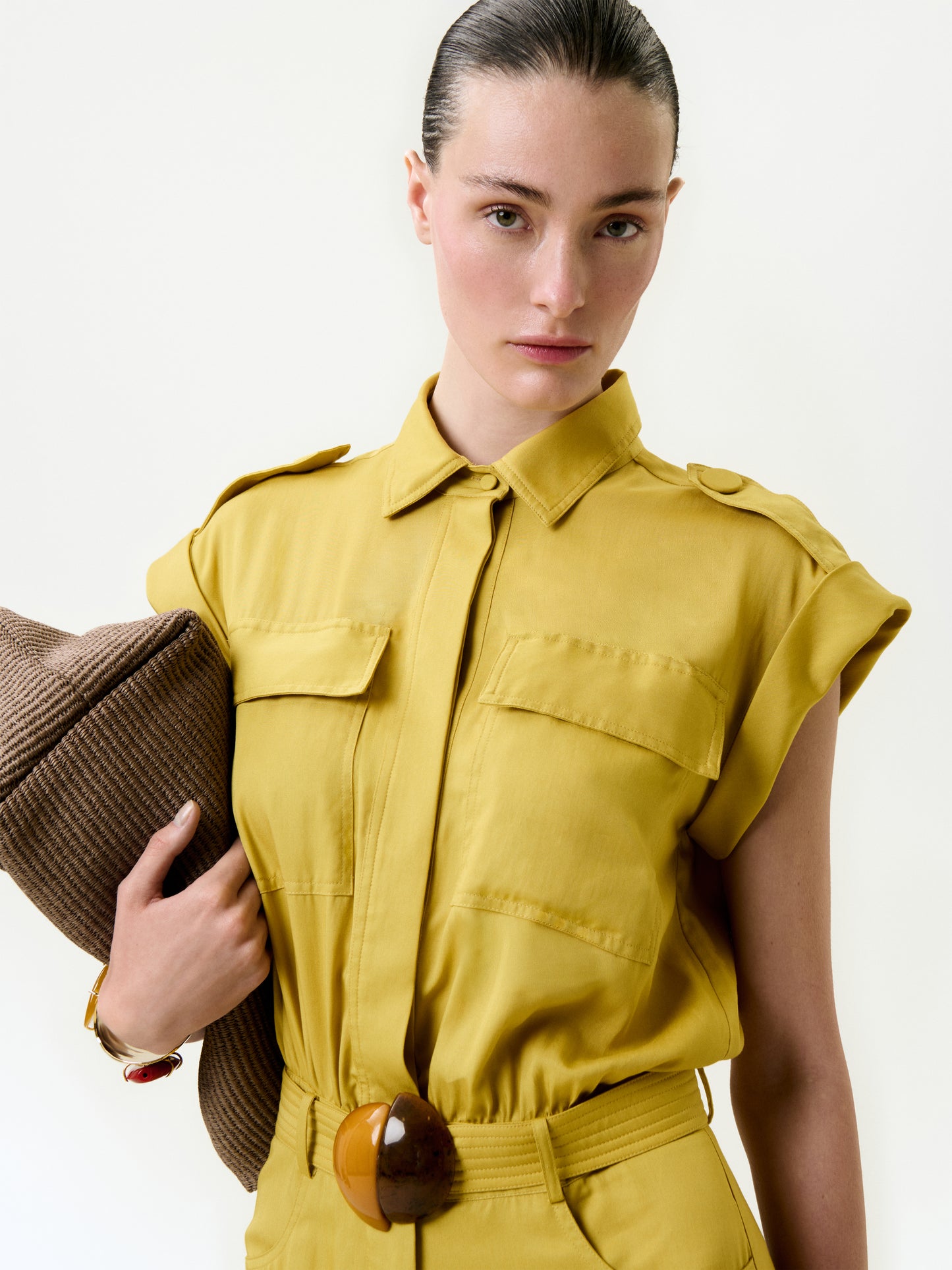 A woman with slicked-back hair and a neutral expression stands against a plain light background, wearing the Sabrina Jumpsuit—a mustard yellow, short-sleeve style in soft Italian fabric with chest pockets and a wide belt—holding a brown hat under her arm.
