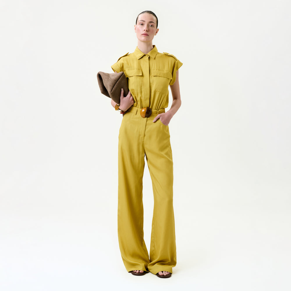 A person stands against a plain white background wearing the Sabrina Jumpsuit in mustard yellow, featuring wide legs and short sleeves, paired with a large brown clutch and brown sandals.