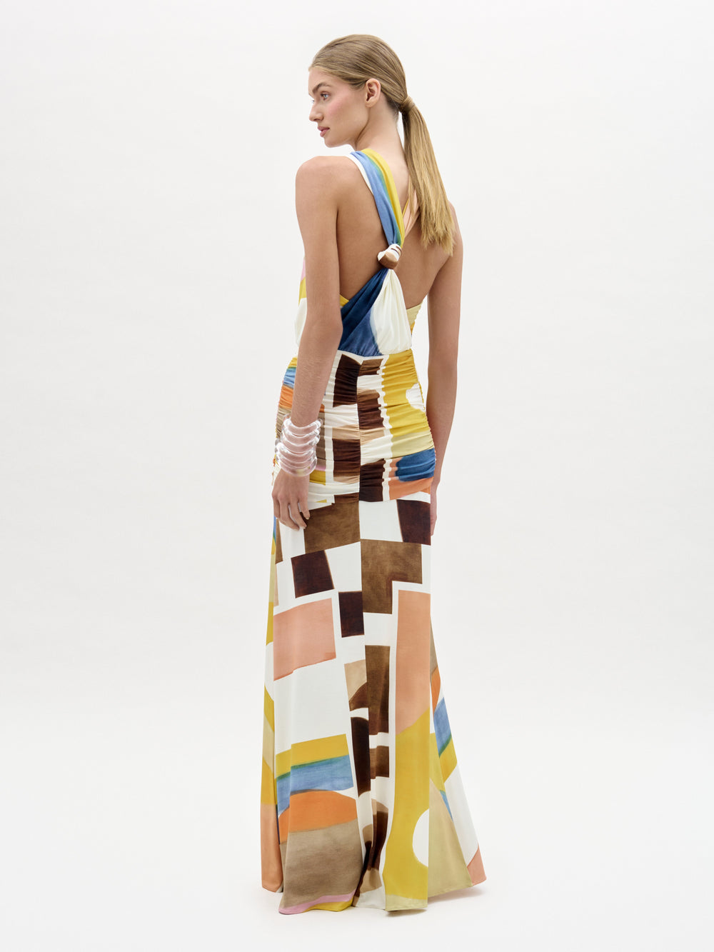 A woman stands against a white background wearing the Jedda Dress, a colorful abstract-patterned maxi with a halter neck, ruched details, and strappy sandals.