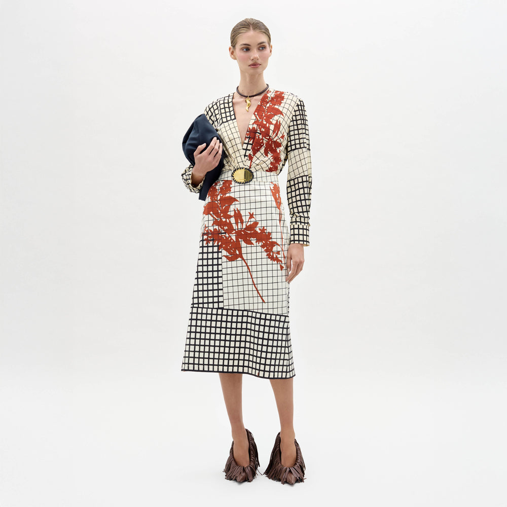 A woman stands against a white background wearing the Kadira Dress, a checked dress with red leaf prints, paired with a black clutch and brown fringed shoes.